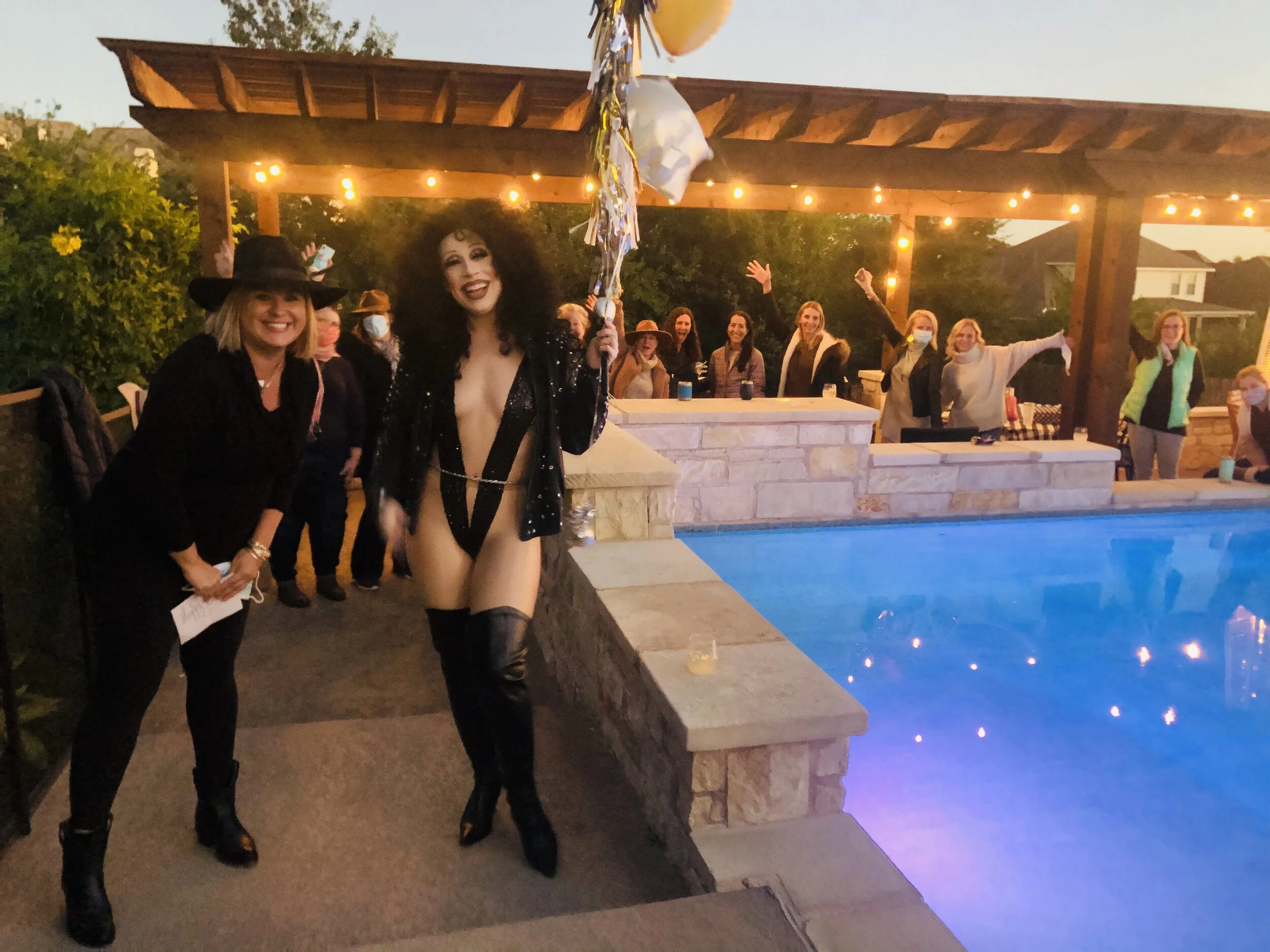 A woman dressed as Cher with curly black hair, wearing a black bodysuit, thigh-high boots, and a blazer, holding balloons, stands by a swimming pool where a party is taking place, with other women in the background celebrating.