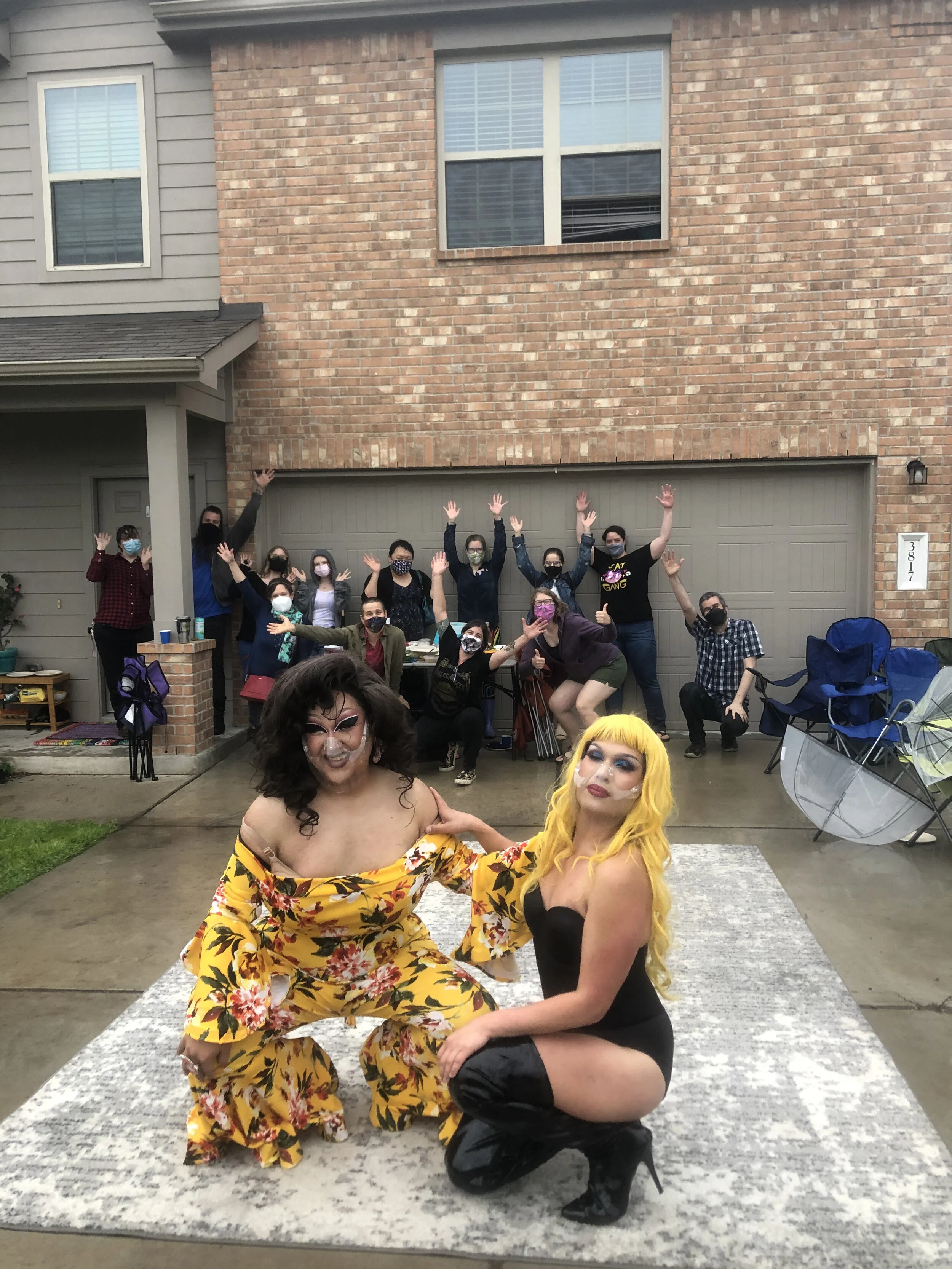 Two drag queens posing in front of a group of people in a residential driveway, with a brick wall and garage in the background. The drag queens are dressed in colorful outfits, one in a floral yellow jumpsuit and the other in a black dress with thigh