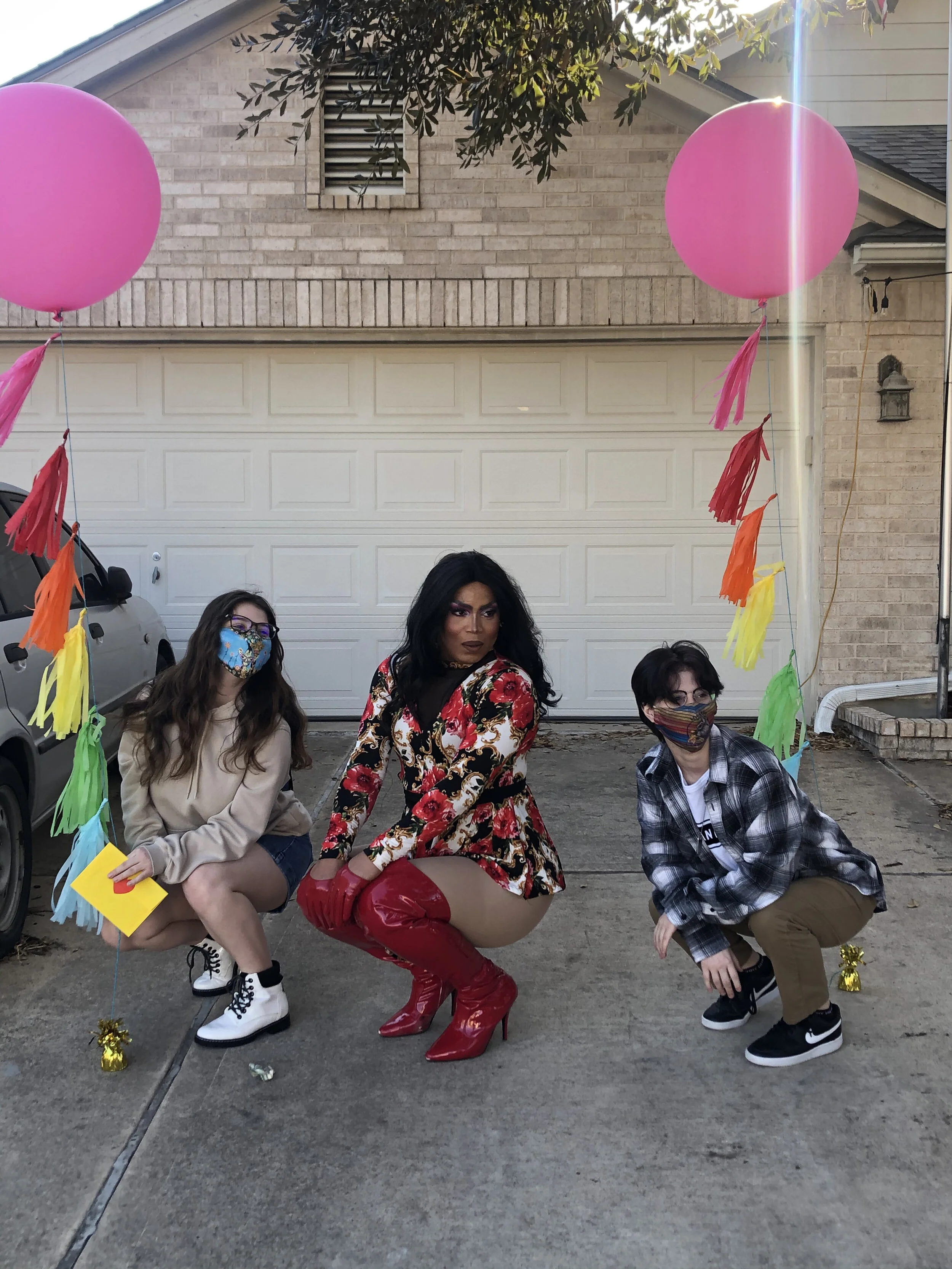 Three people are posing on a driveway decorated with rainbow-colored paper tassels and pink balloons, in front of a garage, with two cars parked nearby. The person in the middle is dressed in a floral dress with red gloves and red high-heeled boots, 