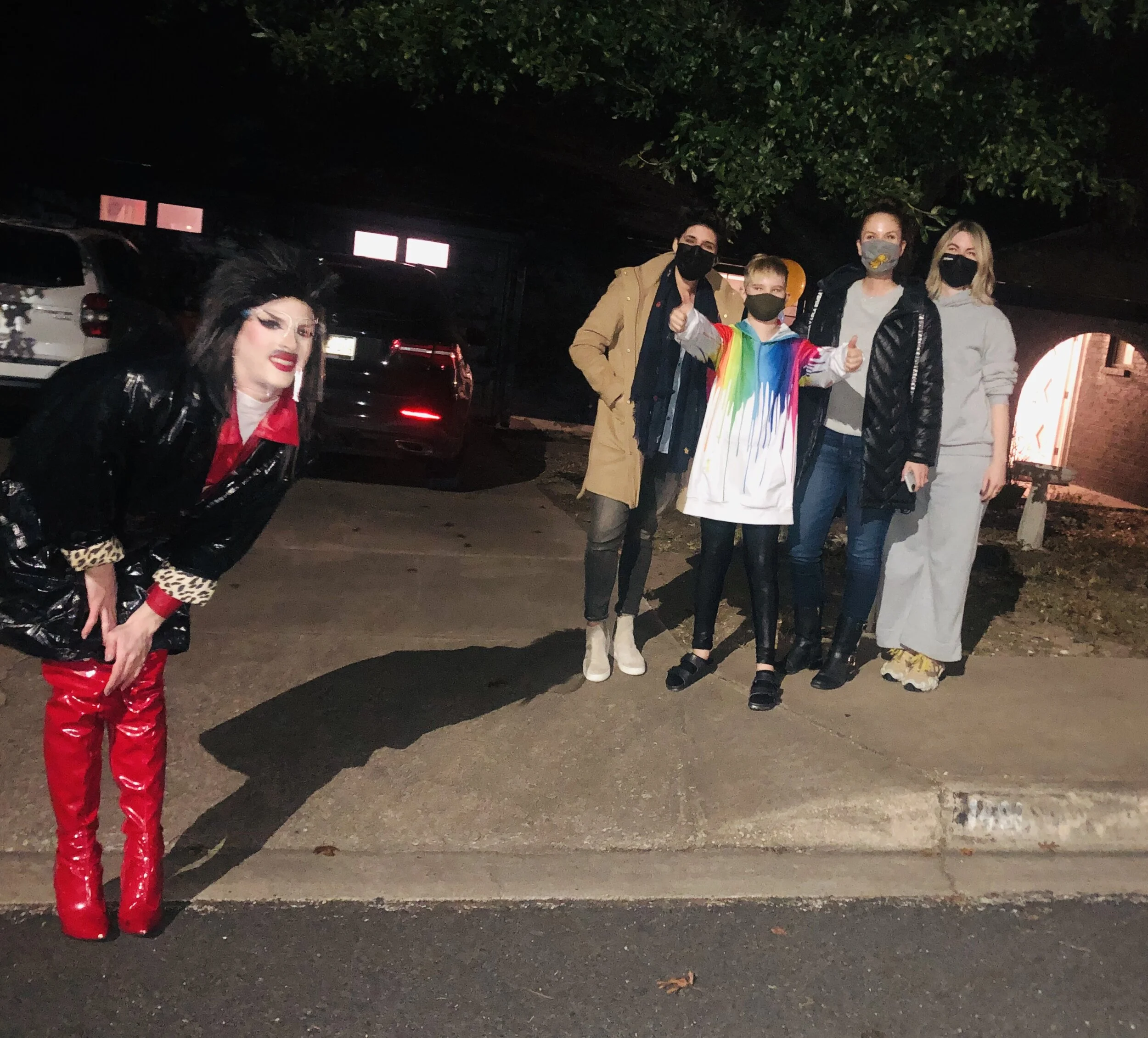 Group of five people, including a drag performer with makeup and shiny red boots, standing outdoors at night on a sidewalk in front of parked cars and dark foliage.