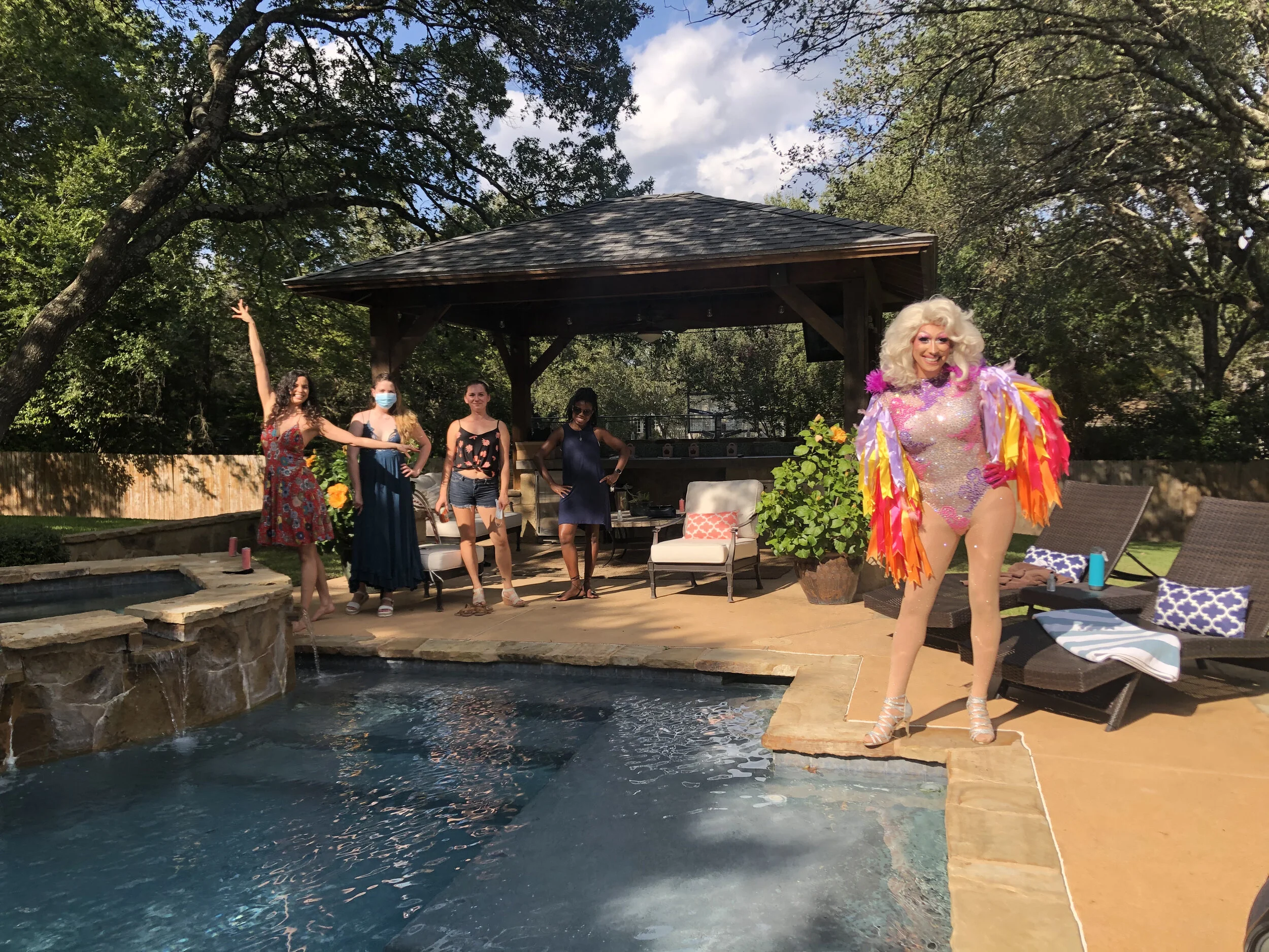 A woman dressed in a colorful, glittery outfit with feathered sleeves posing near a pool, with four other women and a person standing in the background on a patio area in a backyard with trees and a gazebo.