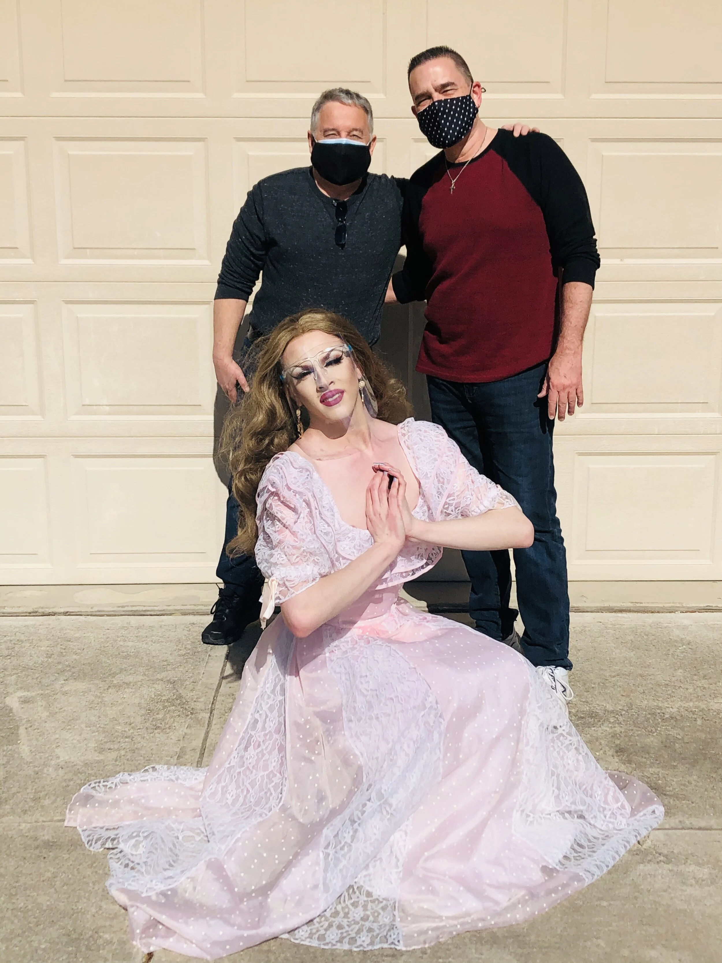 Three people pose in front of a garage door, two men standing behind a seated woman dressed in a pink, lace gown while the men wear black face masks.