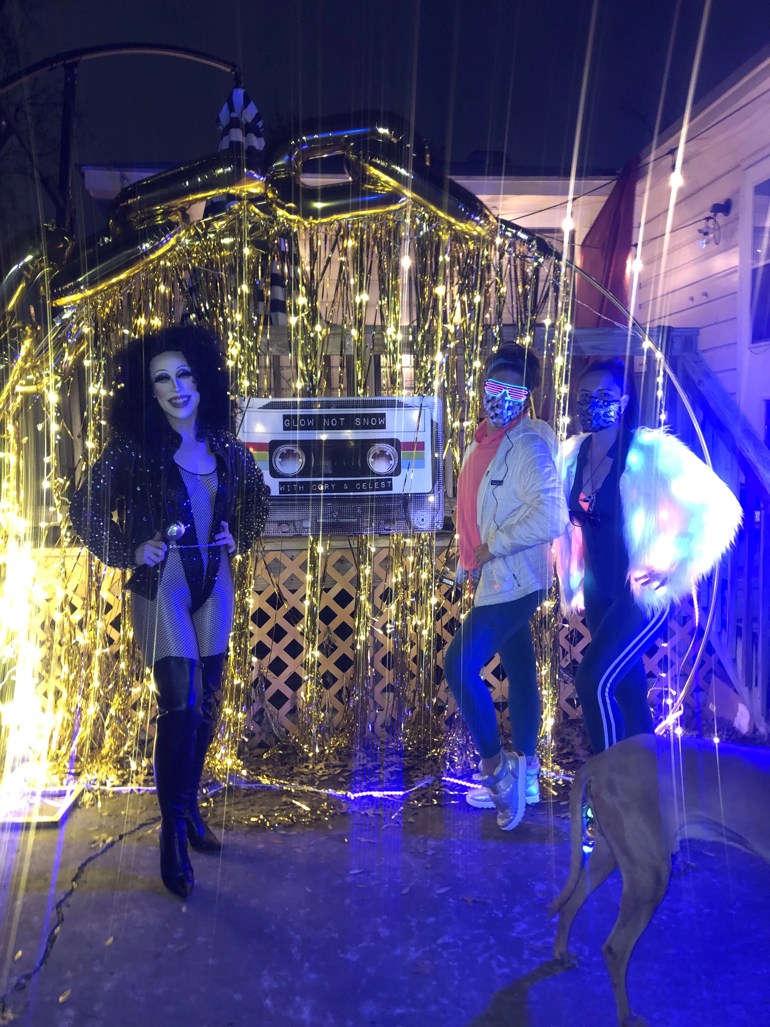 Three people dressed in LED and furry costumes stand inside a decorated enclosure with golden streamers, a cassette tape, and festive lights, possibly at a party or event, with a dog in the foreground.