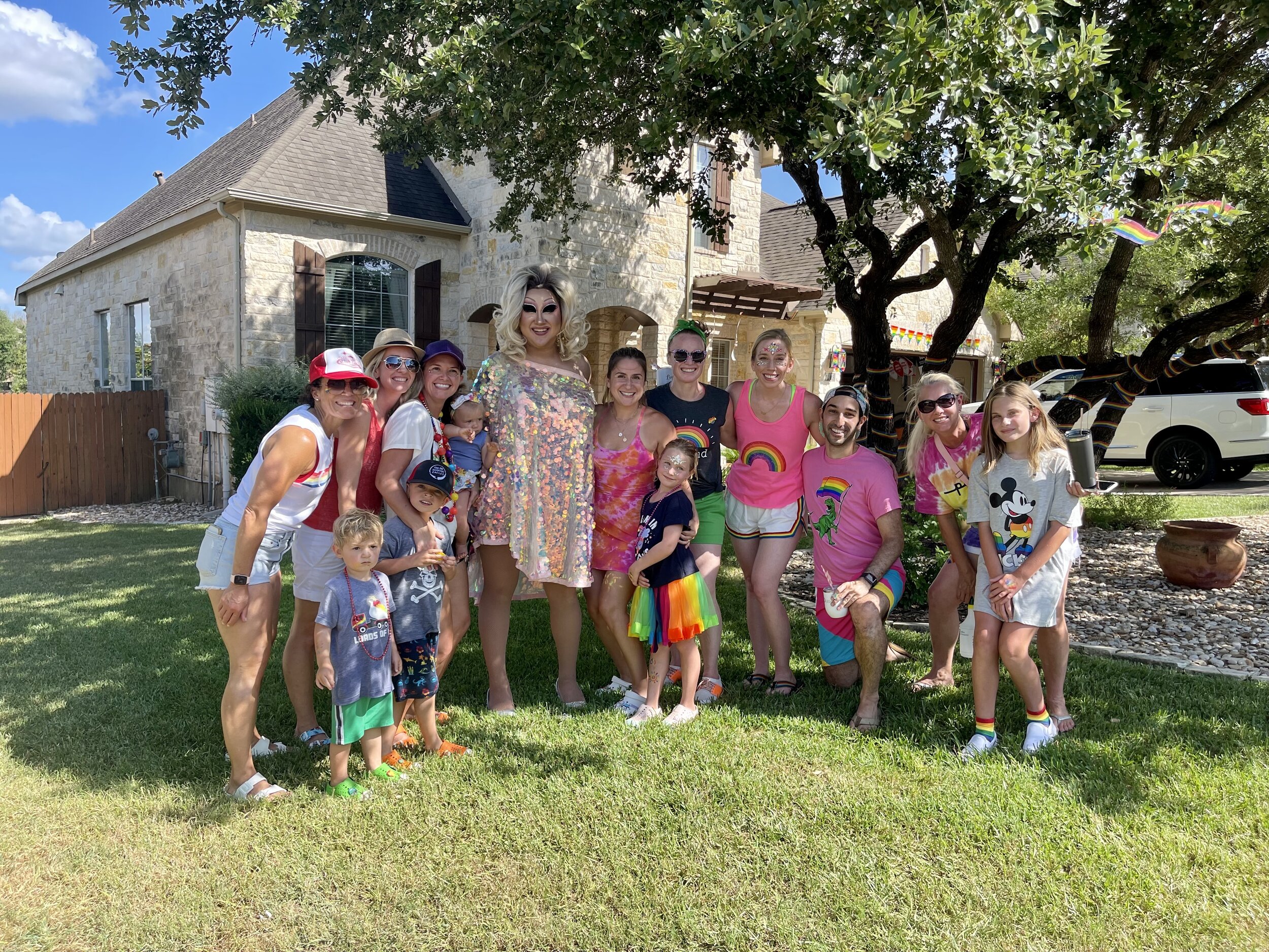 Group of people celebrating Pride outdoors on a sunny day, with rainbow-themed clothing and decorations, including a drag queen, children, and adults, standing on grass in front of a house and a leafy tree.