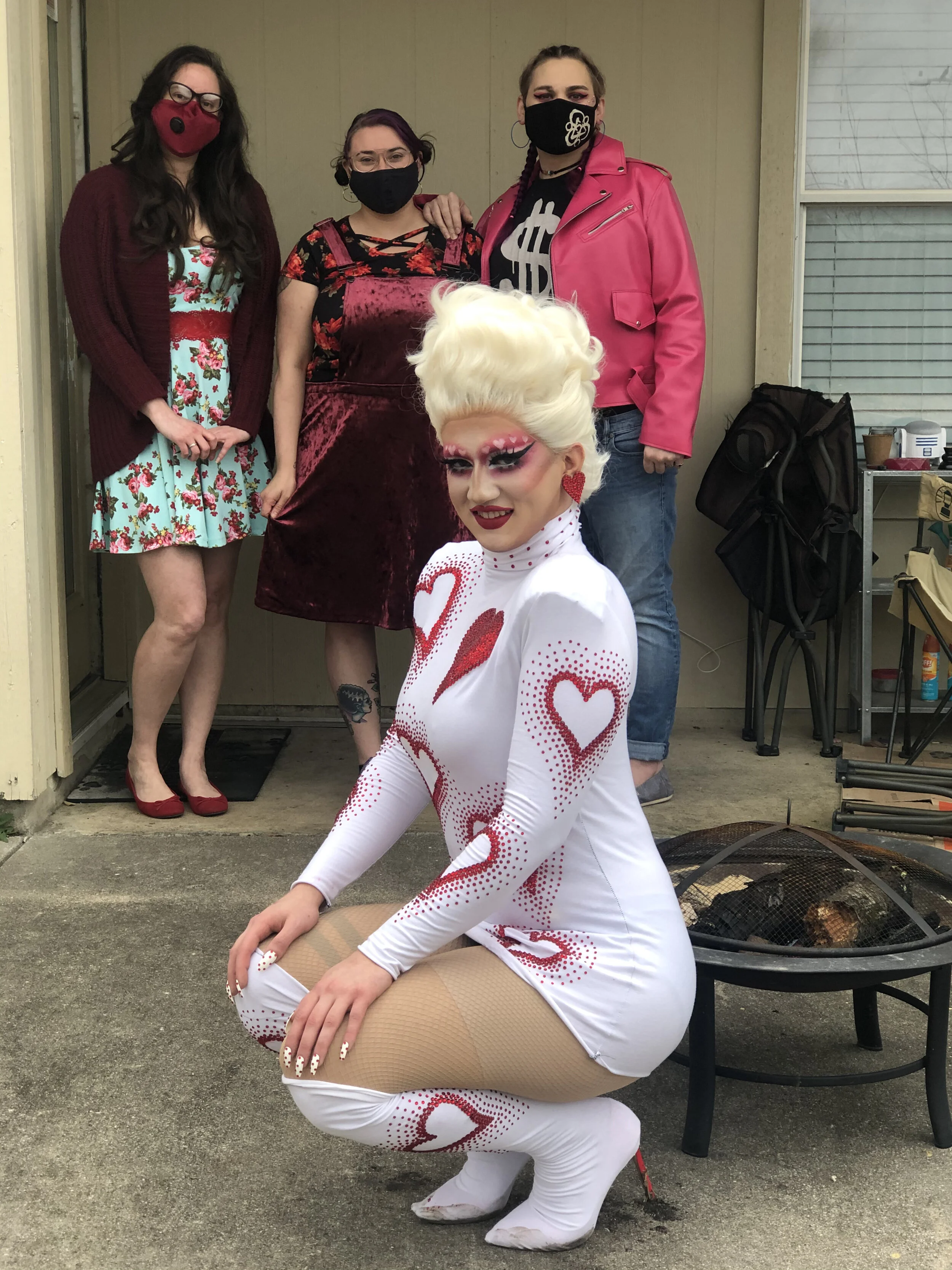 A drag queen kneeling in front of three women wearing masks, with a fire pit behind her.