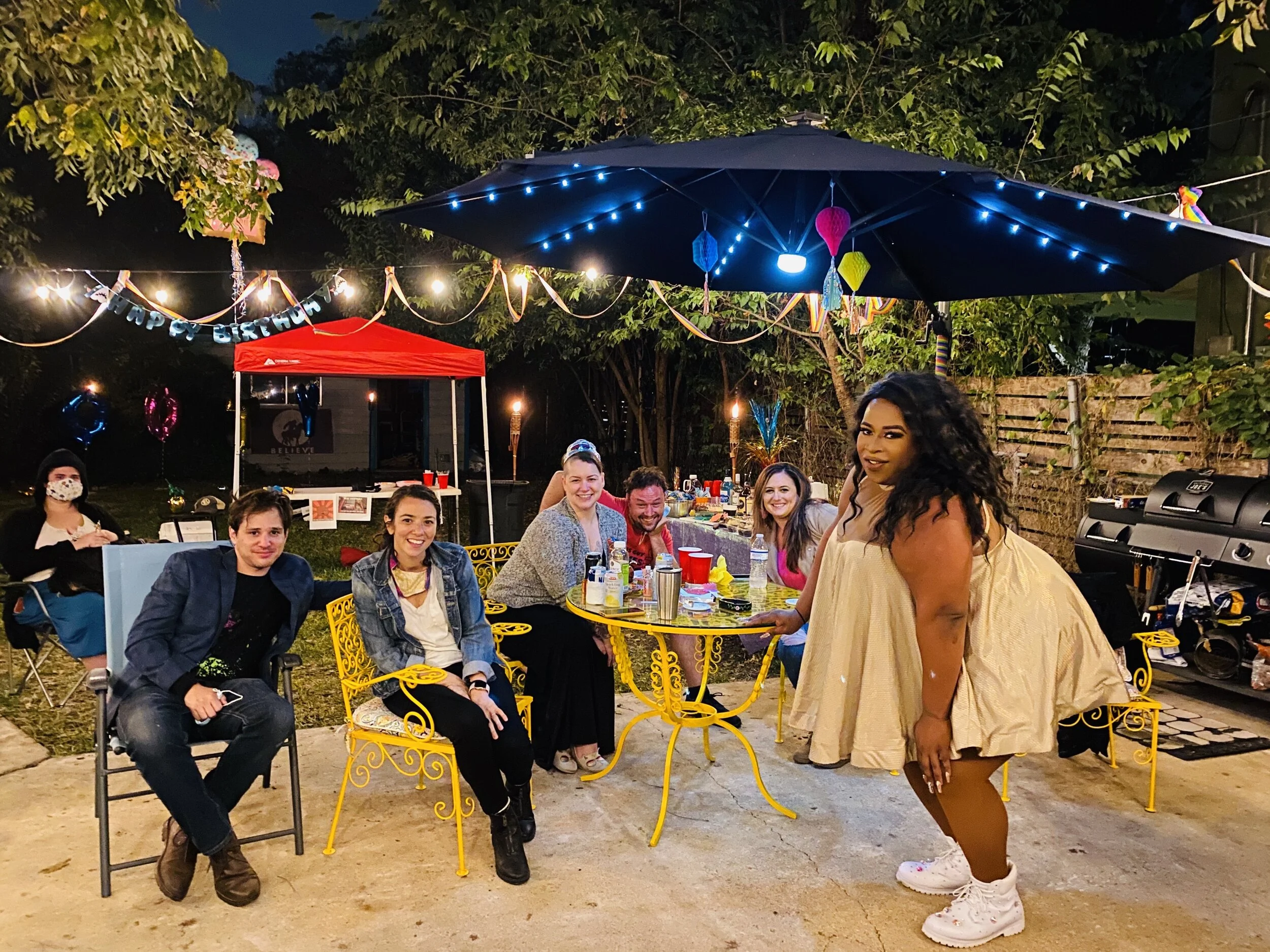 Group of people celebrating birthday outdoors at night with string lights, balloons, and a table with food and drinks, under a large umbrella and a red canopy, in a backyard setting.
