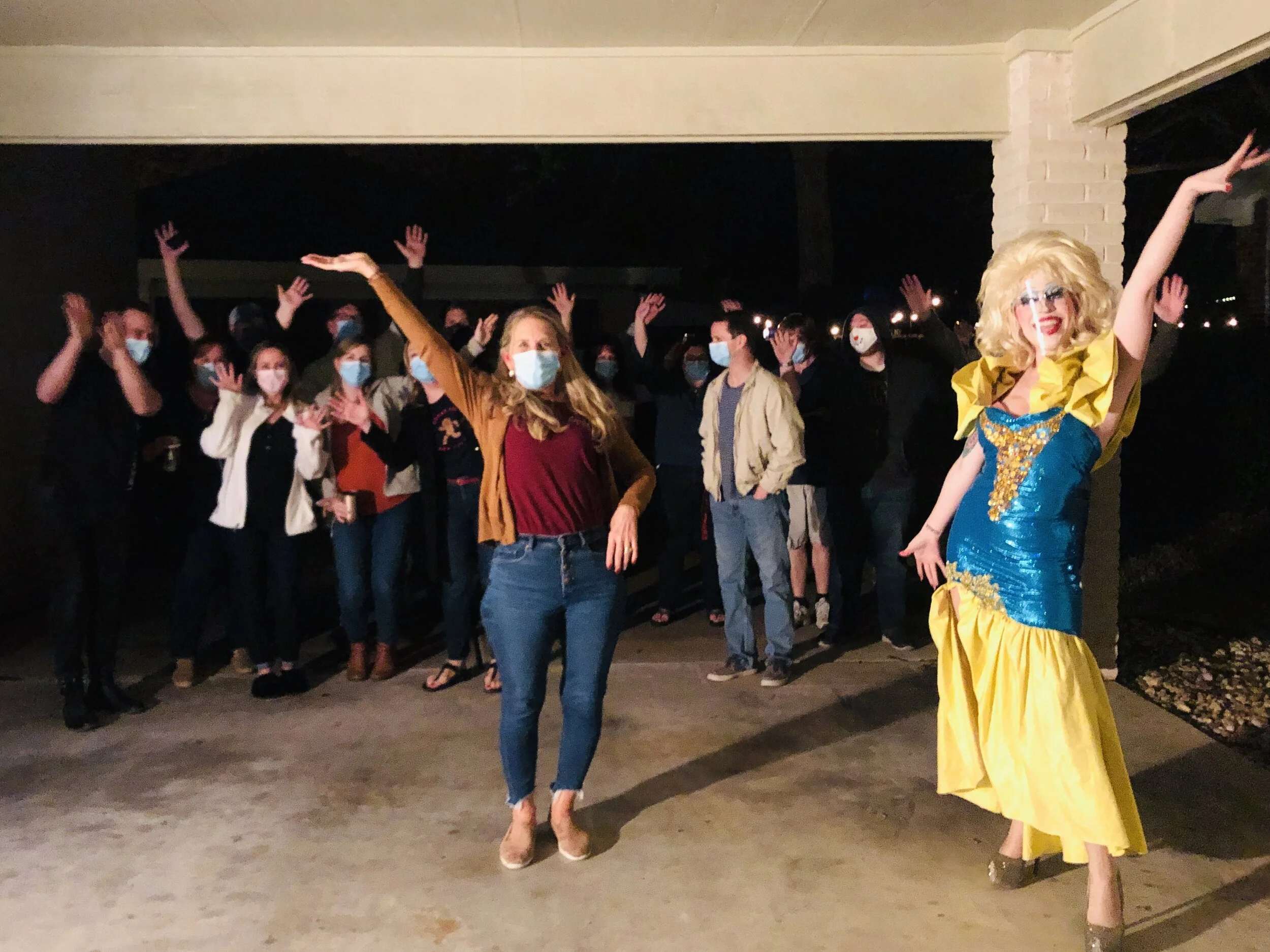 A group of people wearing face masks at night, with two individuals in the foreground: a woman in casual clothing and another dressed as a drag queen in a colorful costume.