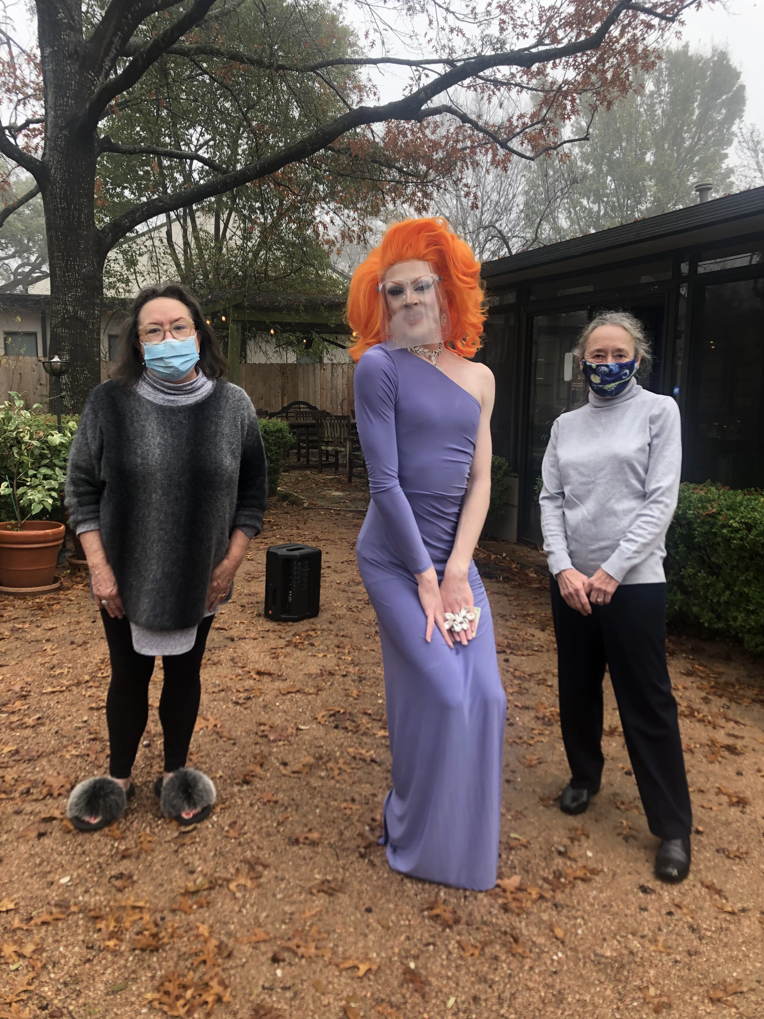 Three women standing outdoors on a cloudy day, wearing masks. The woman in the middle has bright orange hair and is dressed in a purple gown with a one-shoulder long sleeve. The woman on the left has dark hair, glasses, and is wearing a gray sweater,