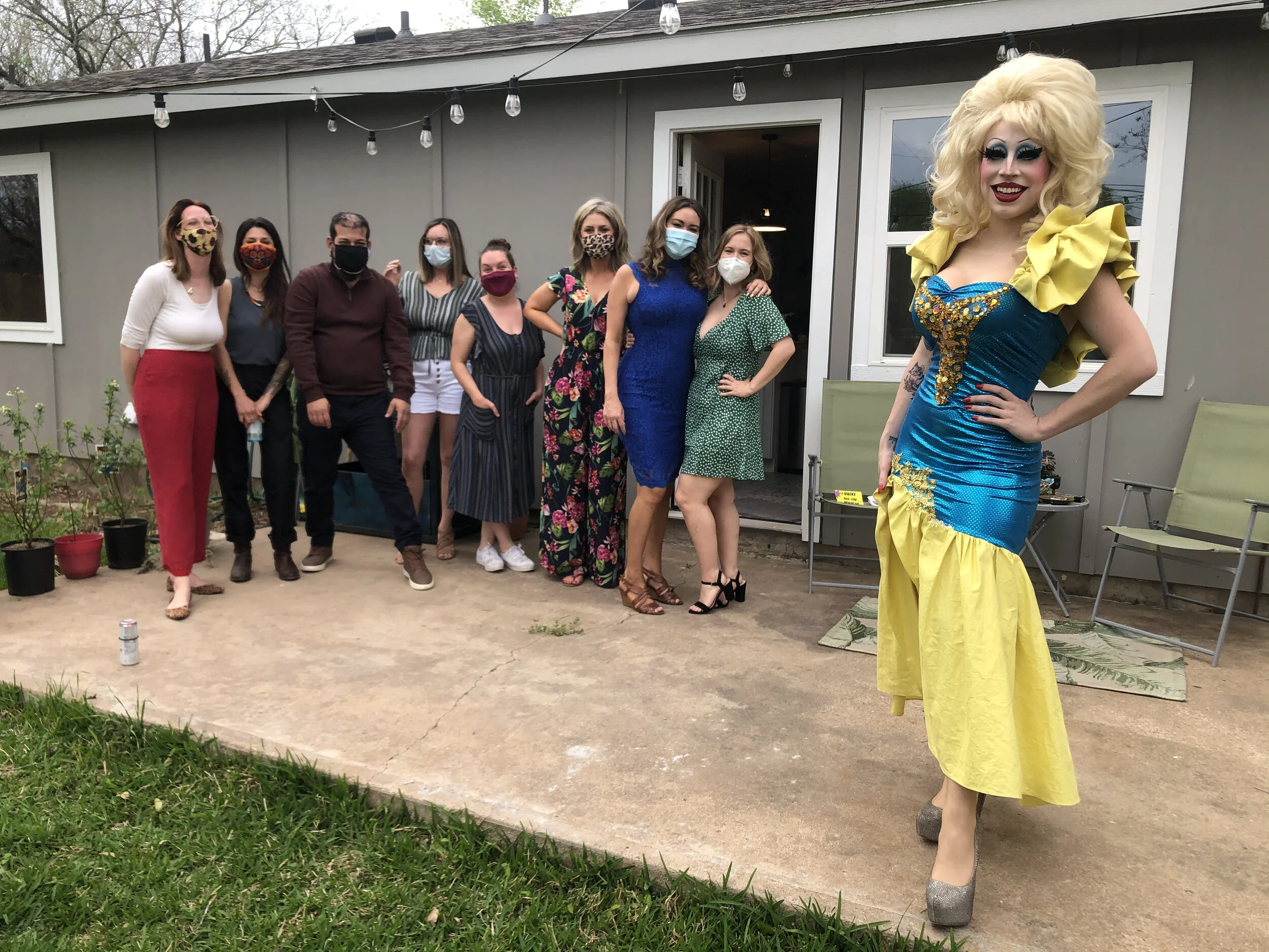 A group of nine women standing outside on a patio with one dressed as a drag queen in a colorful dress and high heels. All individuals are wearing face masks.