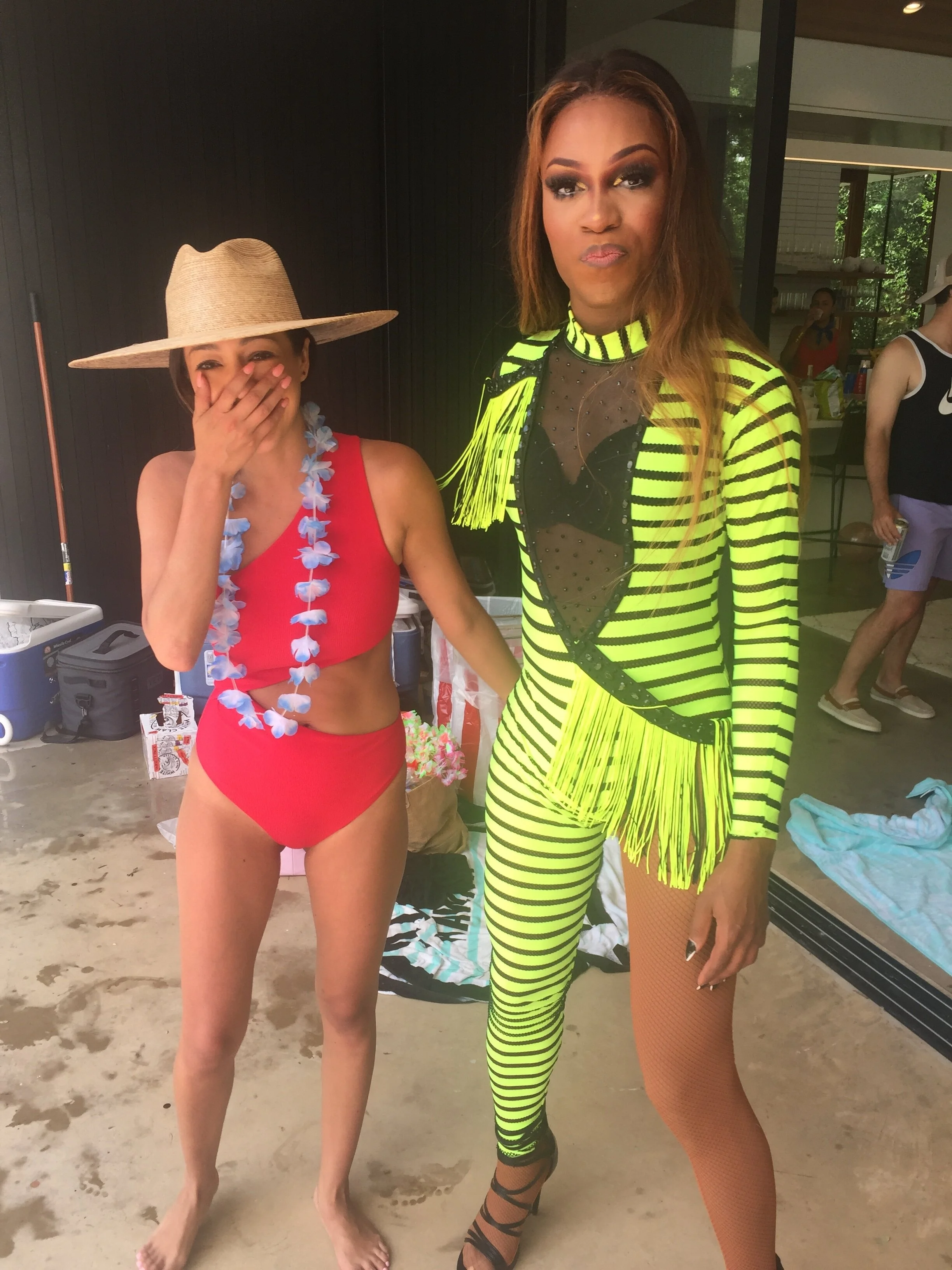 Two women in colorful outfits, one in a red swimsuit with a straw hat and lei, and the other in a neon yellow and black striped costume with fishnet stockings, standing indoors near a glass door.