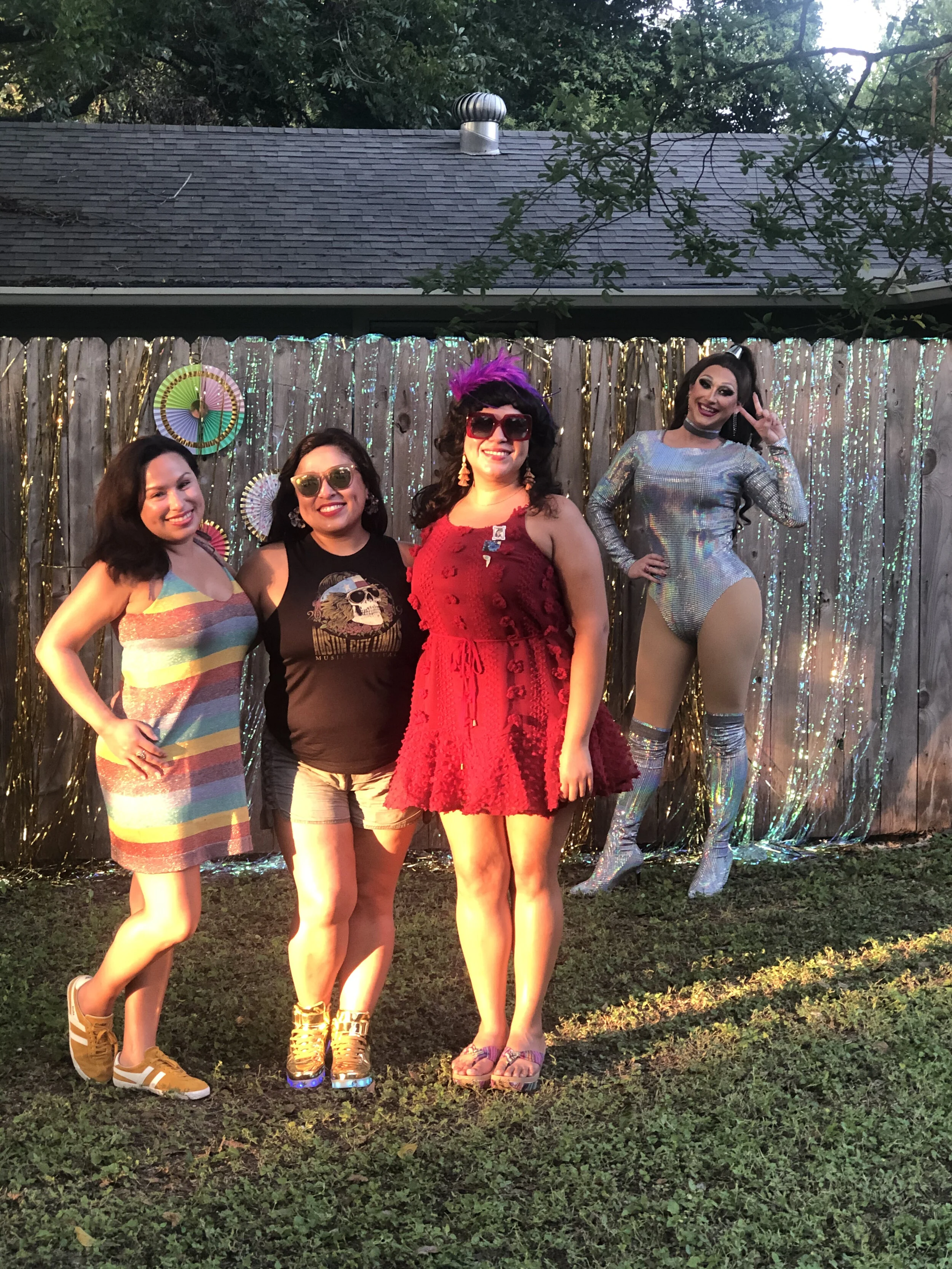 Four women posing in front of a decorated outdoor backdrop with metallic streamers and fans. Three women are standing together, smiling, with one wearing a rainbow-colored dress and sneakers, another in a Coca-Cola shirt and shorts, and the third in 