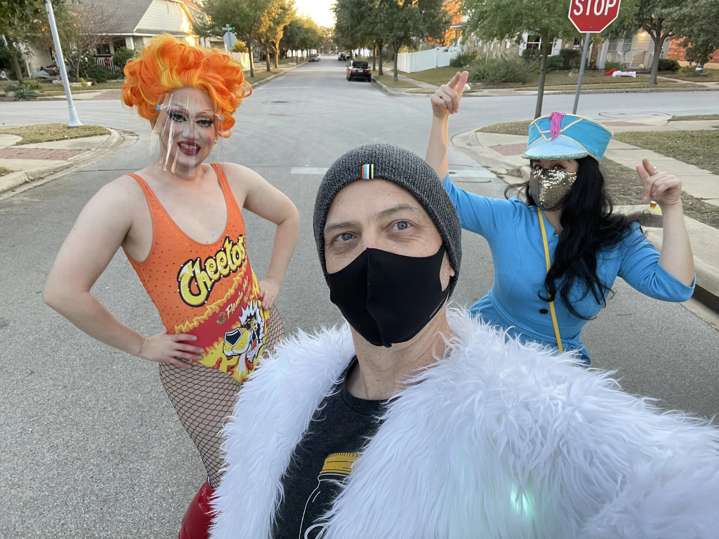 Three people dressed in colorful costumes taking a selfie outdoors in a residential neighborhood. A person in the foreground wears a gray beanie and a black face mask, with a white fluffy jacket. To the left, a person with bright orange hair, makeup,