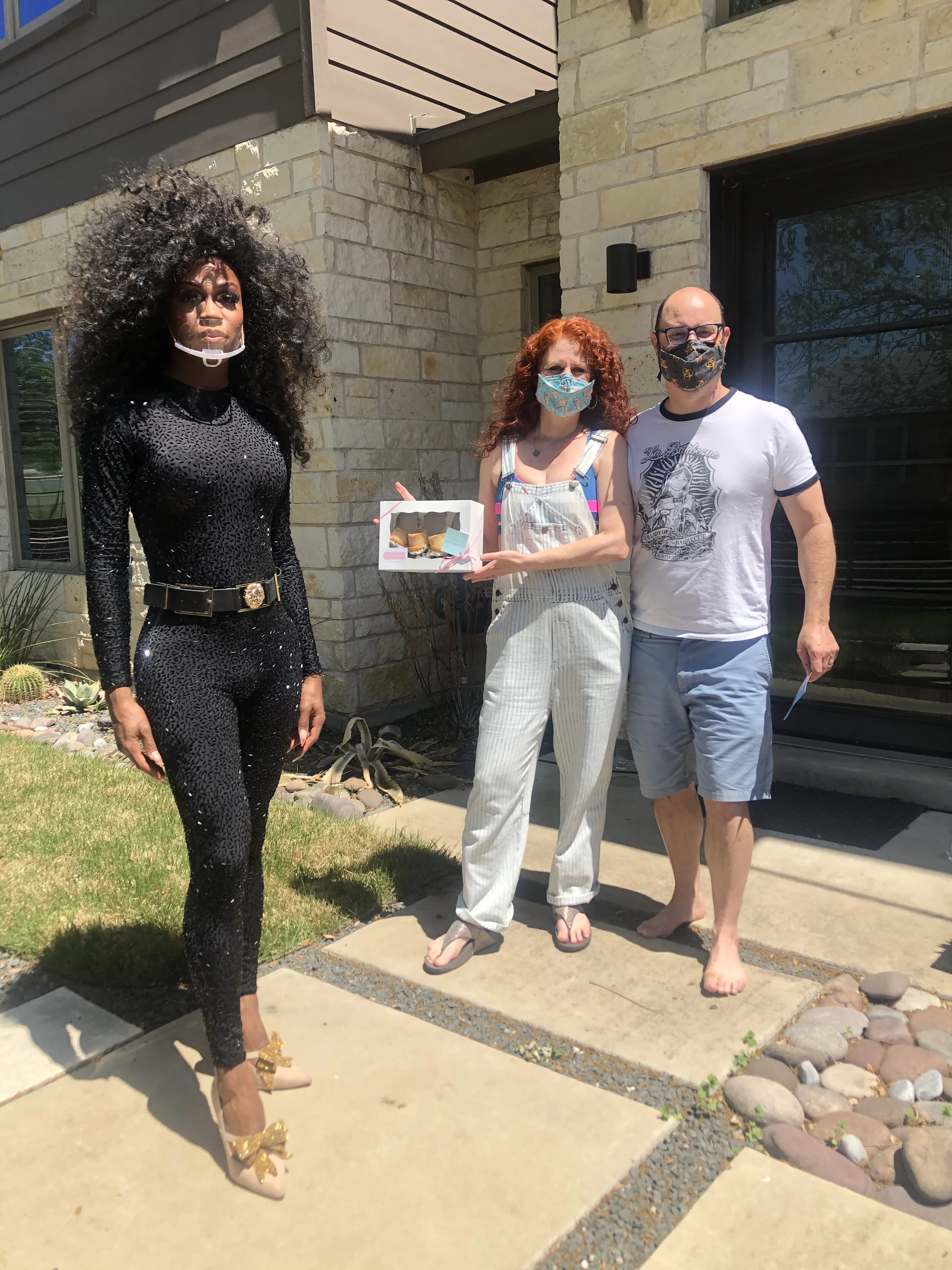 Three people standing outside in front of a house, with one woman in a black sequined outfit and high heels, another woman holding a small box of pastries, and a man, all wearing masks.
