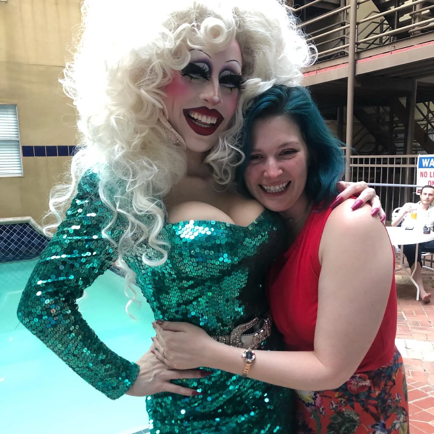 Two women smiling and hugging outdoors; one dressed as a drag queen with a curly platinum blonde wig, dramatic makeup, and a shiny green sequined dress; the other with shoulder-length teal hair, wearing a red sleeveless top.