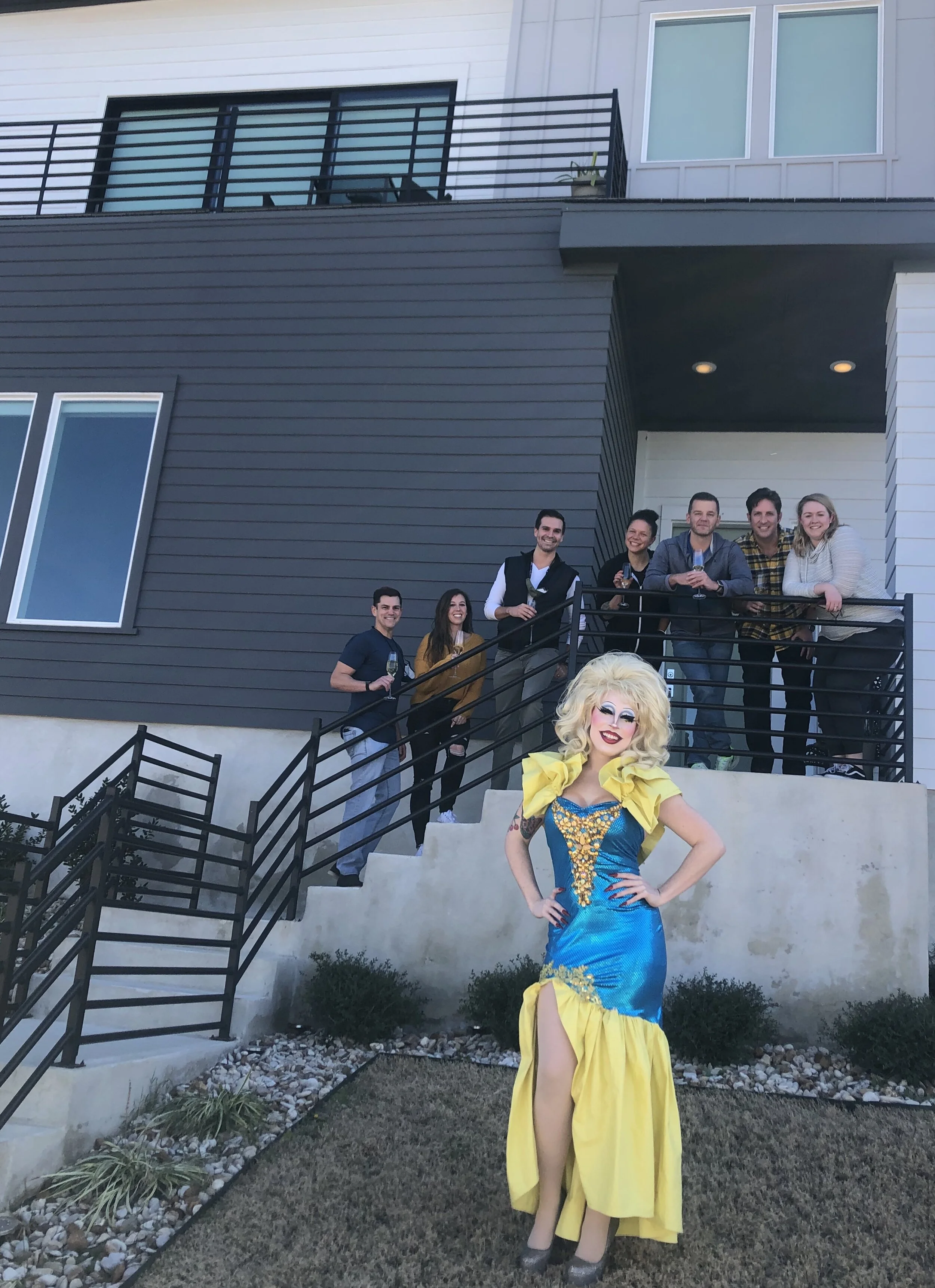 A group of eight people standing on a porch with black metal railings, including a person dressed as a drag queen in a colorful yellow and blue dress with blonde curly hair, posing in front of a modern, multi-story residential building.