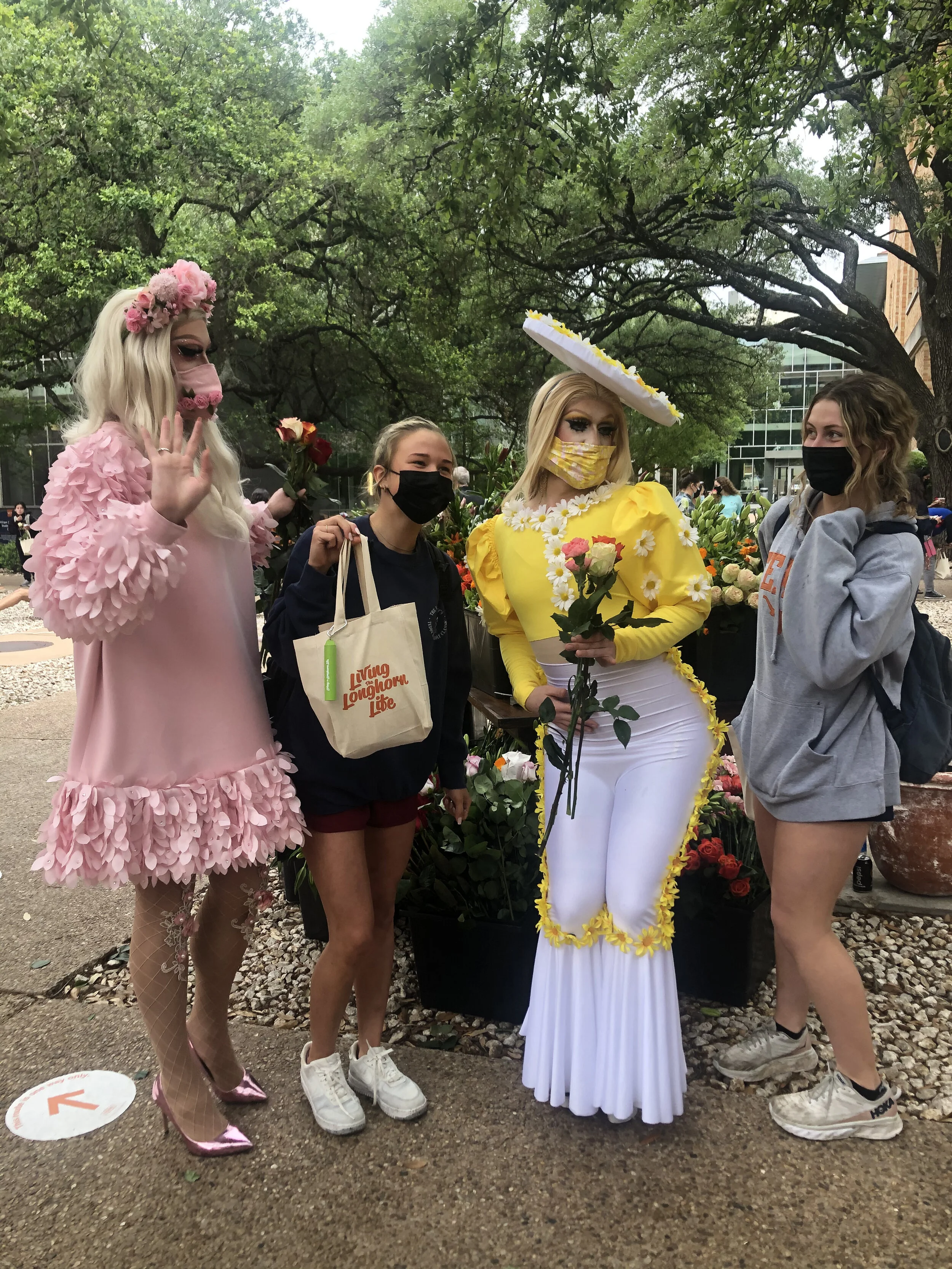 Four women standing outdoors with trees in the background, one dressed in a Peep-themed costume with pink fluff, one holding a rose, another dressed in an Easter-themed yellow and white dress with a wide-brimmed hat, and the last in a gray hoodie. Al