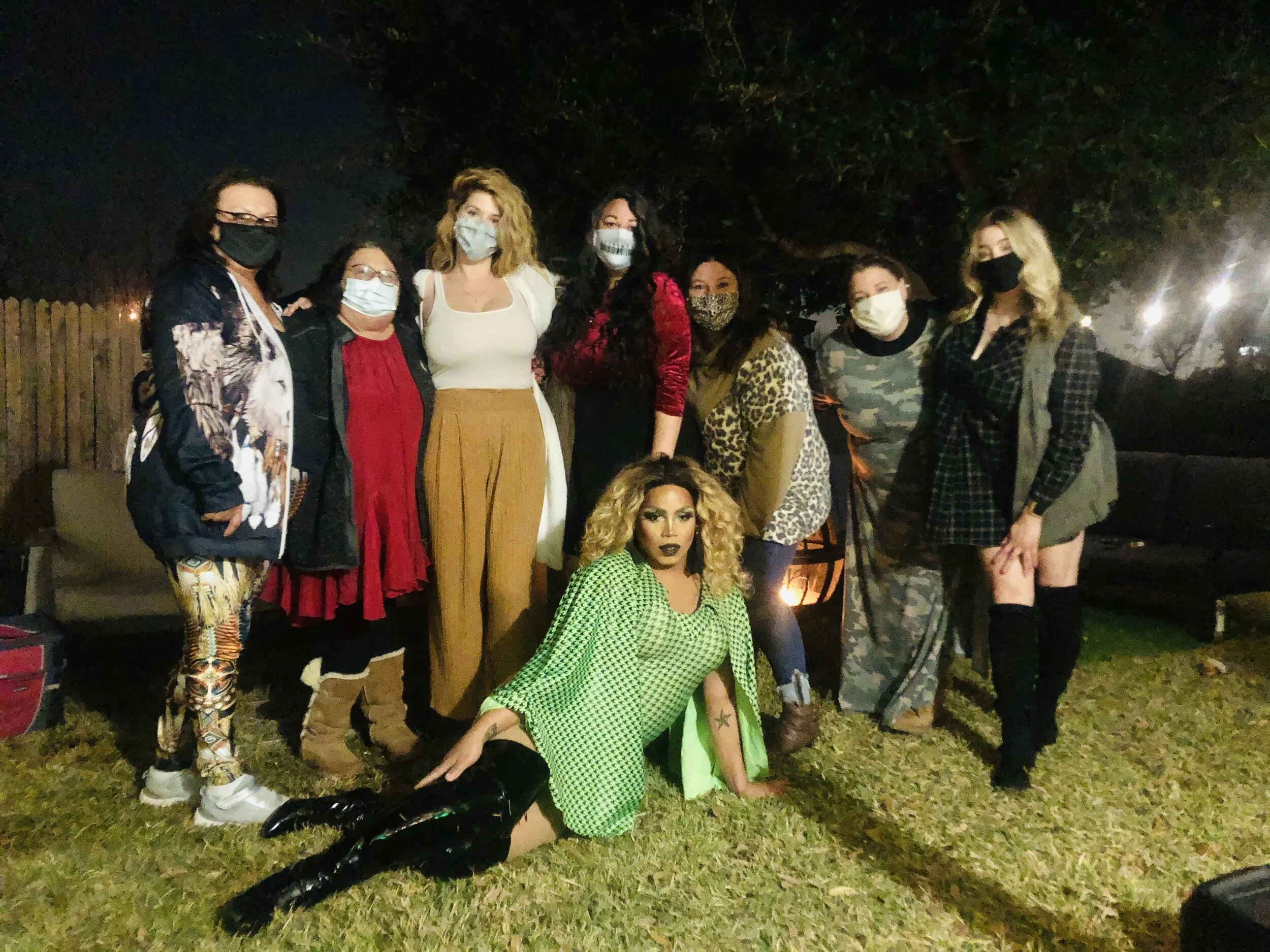 Group of nine women outdoors at night wearing masks, with one sitting on the grass in front.