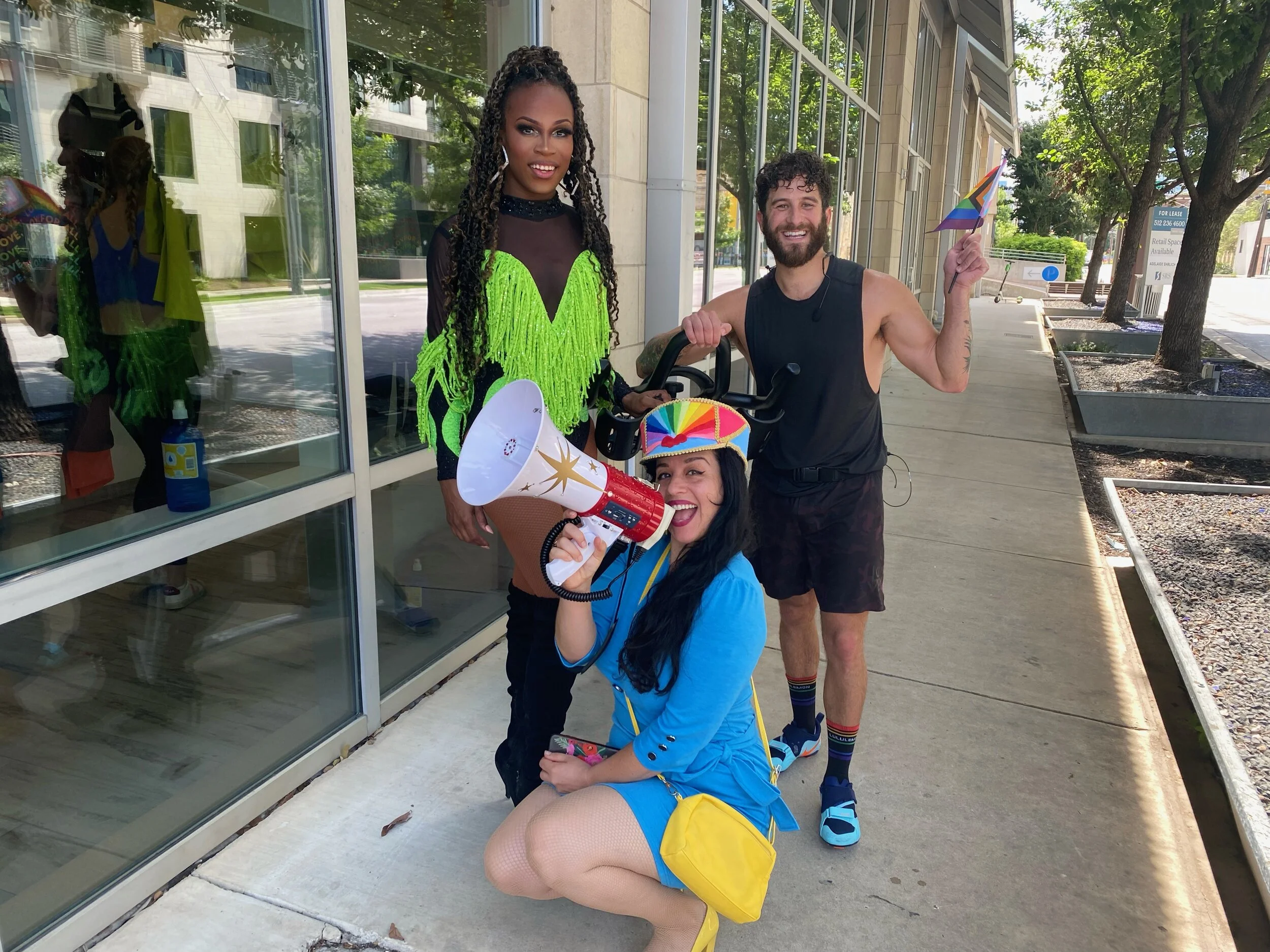 Three people celebrating pride parade, one woman crouching with megaphone, one woman standing in neon green dress, and a man in black tank top holding a rainbow pride flag outside a building with glass windows, trees and sidewalk in background.