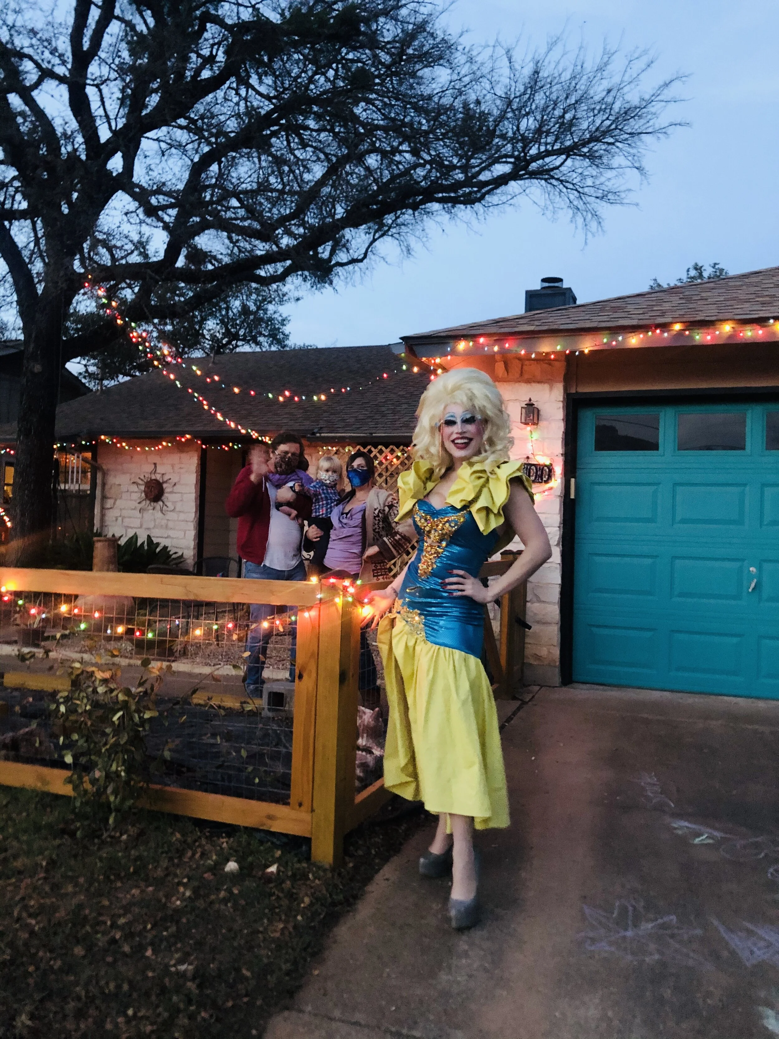 A drag queen in a vibrant blue and yellow dress poses in front of a house decorated with holiday string lights. Several people, some holding children, stand behind a wooden fence, also wearing face masks.