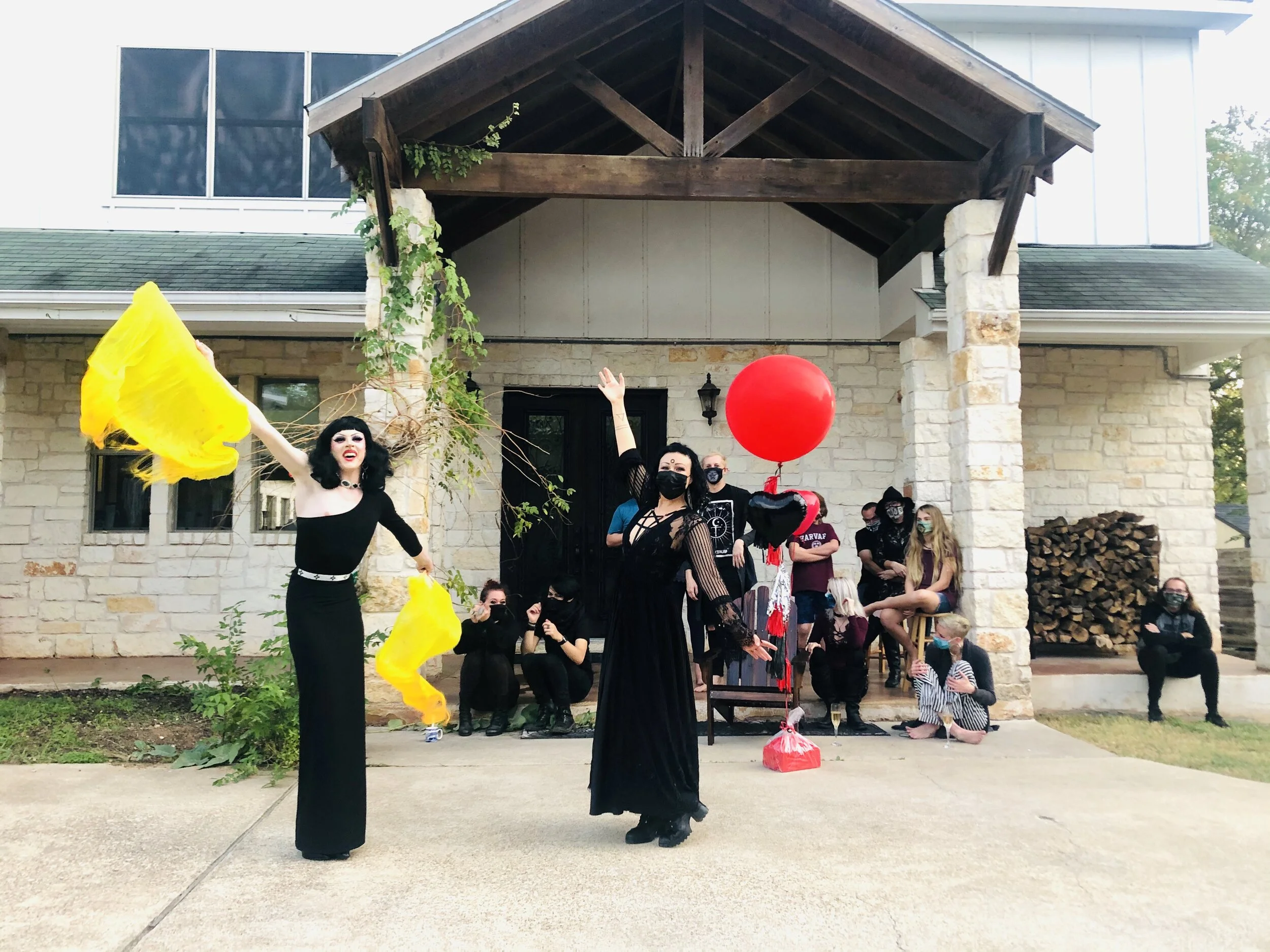 Two women dressed in black costumes dancing in front of a group of people sitting and standing on a porch, with large red and black balloons, and a house with stone and wood exterior in the background.