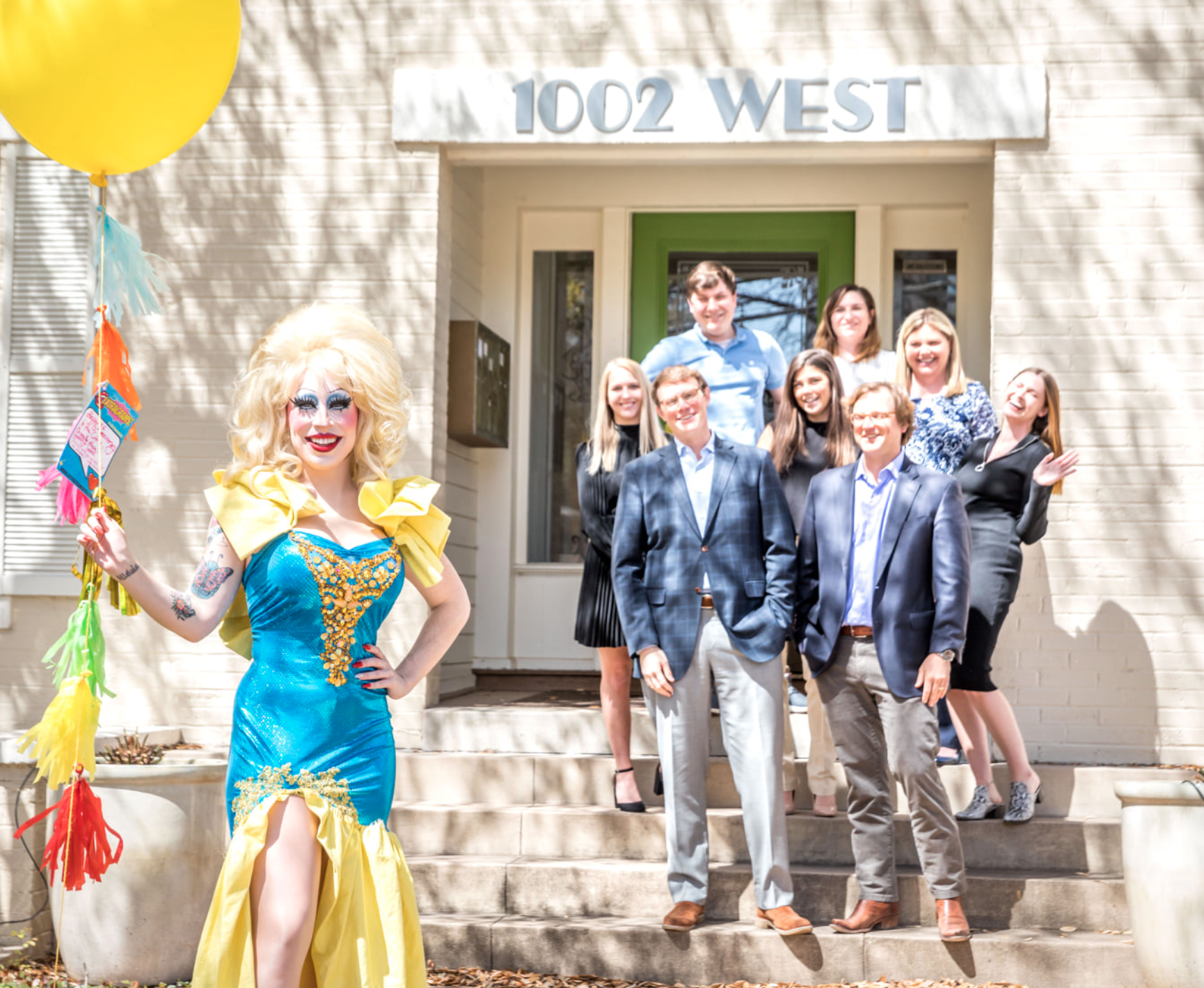 Person dressed as a drag queen standing on front steps holding colorful decorations, with a group of people in business attire smiling behind them outside a building with the address 1002 WEST.