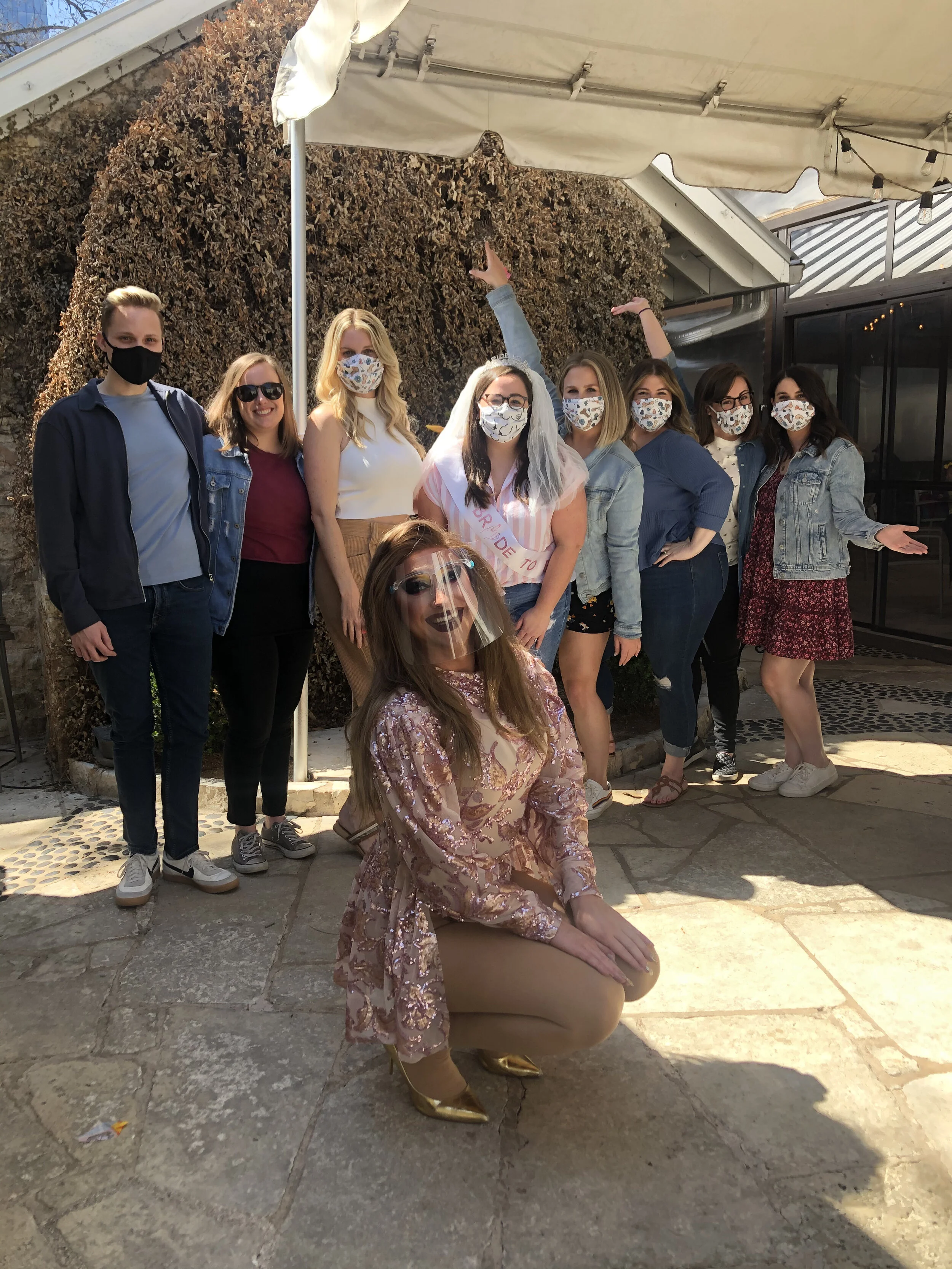 Group of women and young adult in masks outside, celebrating a bridal shower with one woman wearing a sash that reads 'Bride to Be'.