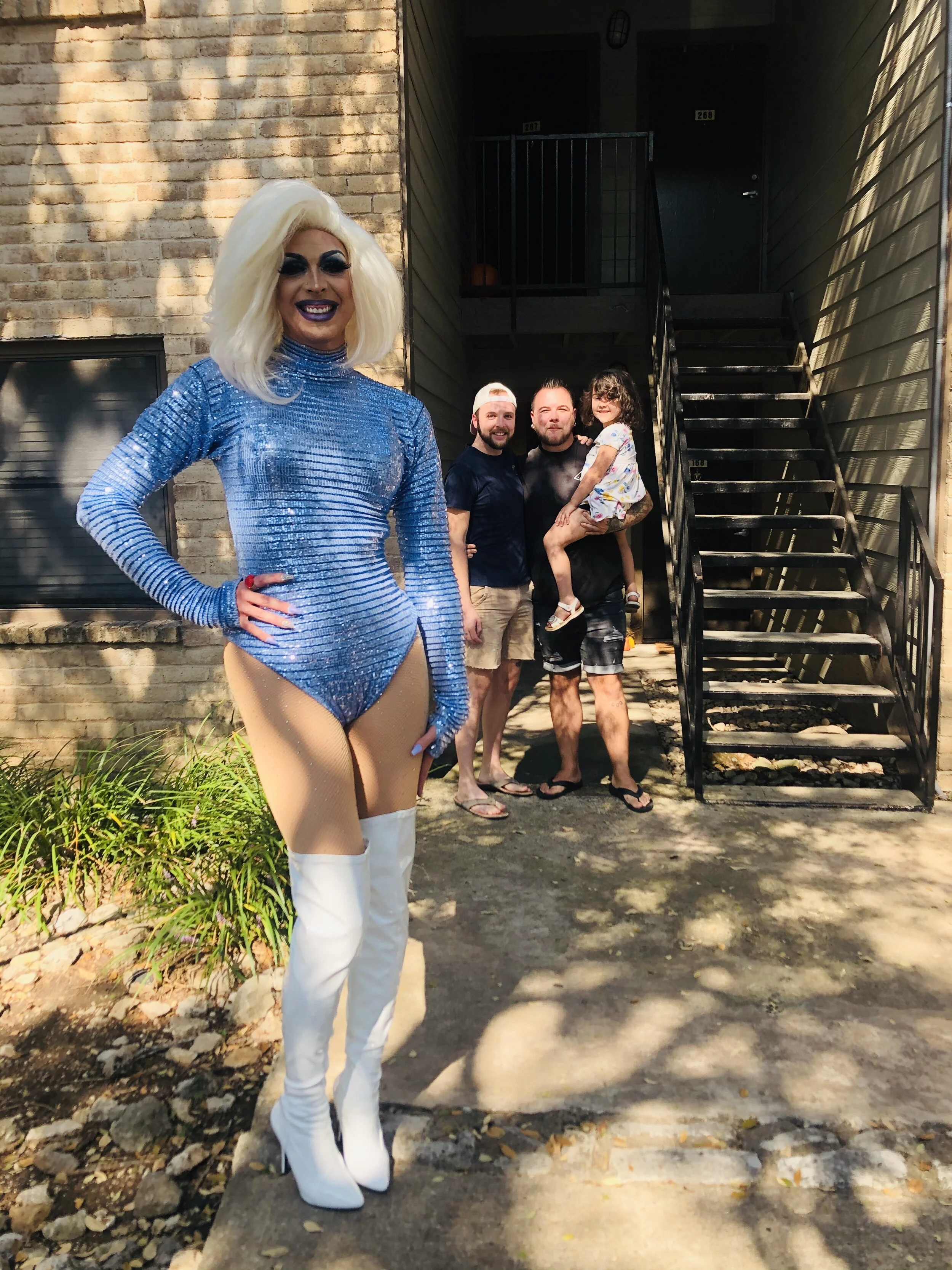 A drag queen with platinum blonde hair, blue makeup, and a sparkling blue outfit with matching gloves and white thigh-high boots standing outdoors in front of a brick building. Three people, two men and a young girl, are standing behind her, smiling.