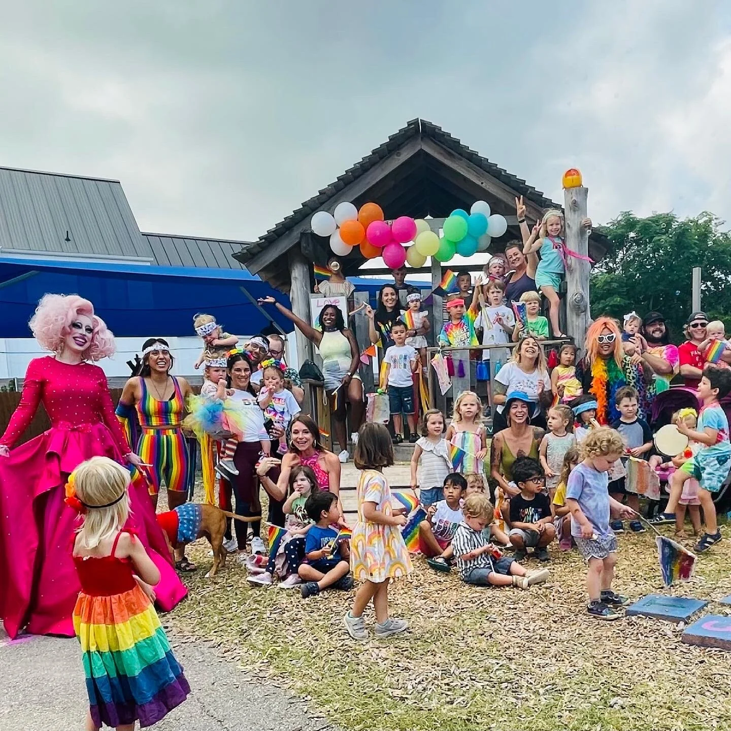 A pride parade or celebration with people in colorful costumes, rainbow accessories, and face paint, gathered on and around a decorated wooden structure with balloons, flags, and rainbow-themed decorations, including children and adults enjoying the 