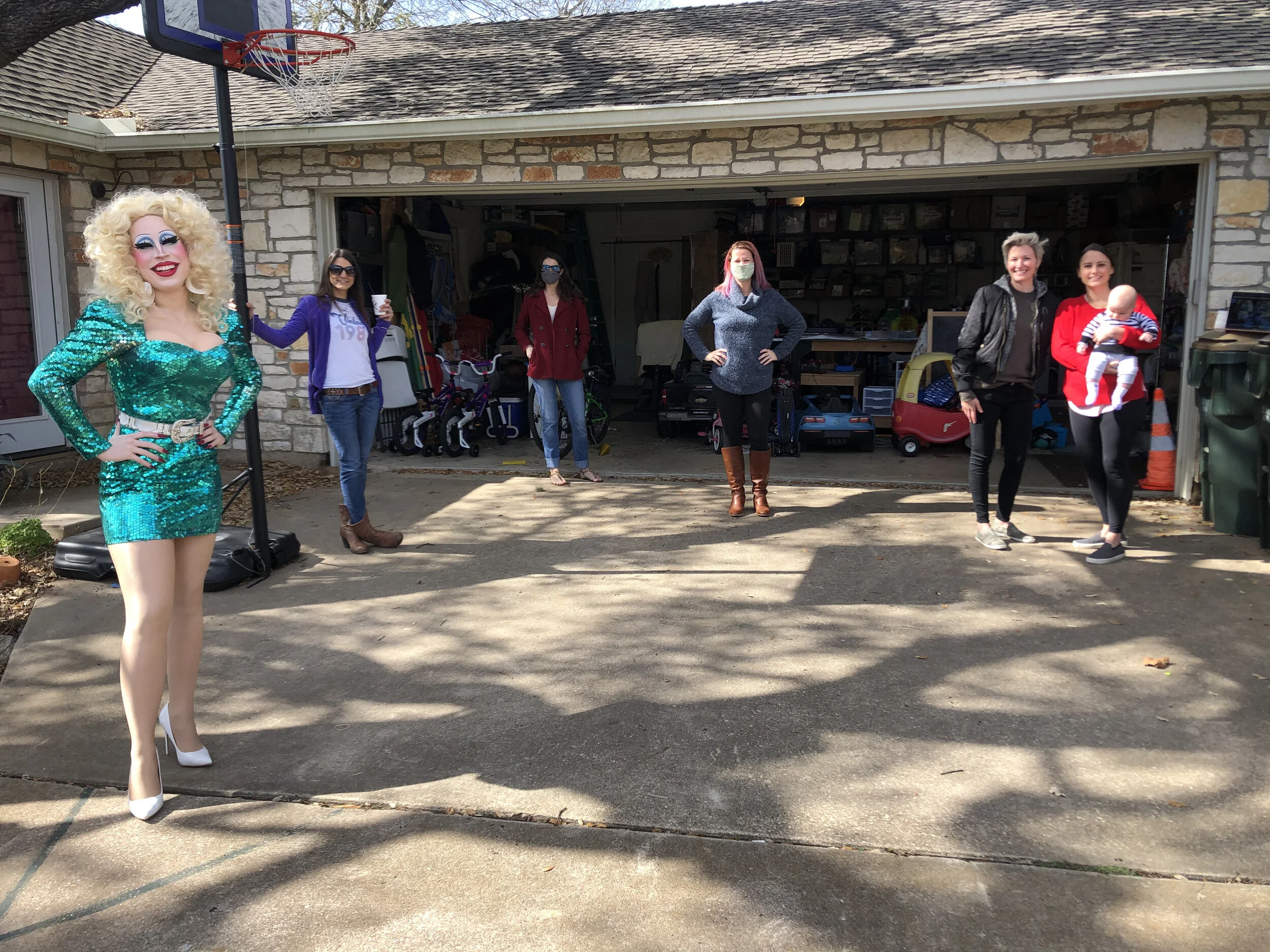 A group of six women, some wearing masks, standing outside a garage. One woman dressed as a drag queen in a sparkling turquoise dress and white heels is in the foreground. The others are in casual clothing, with one holding a baby. There is a basketb