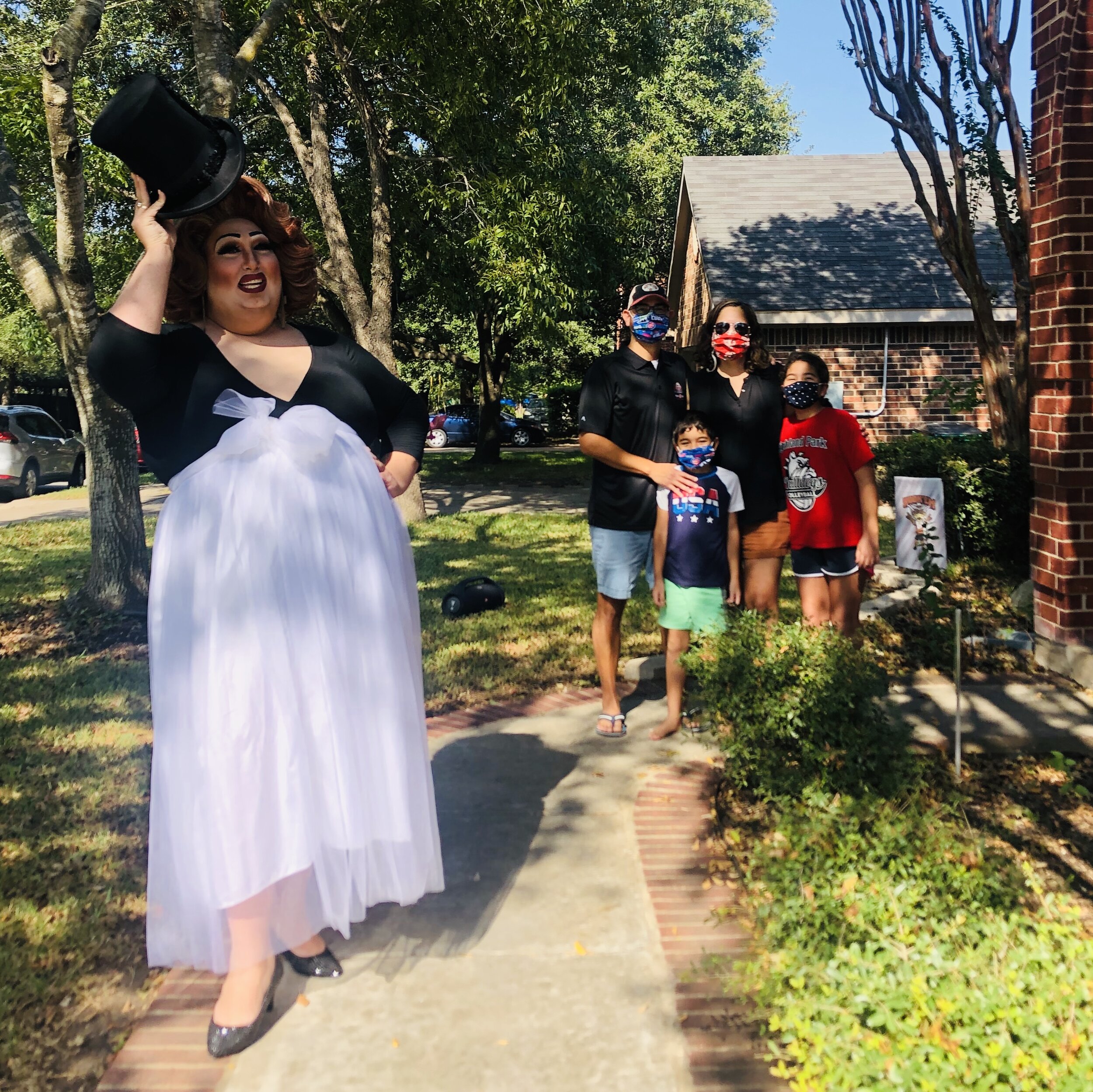 A drag performer in a black top and long white skirt with a black hat lifts the hat while standing on a sidewalk near a family posing for a photo outdoors during daytime.
