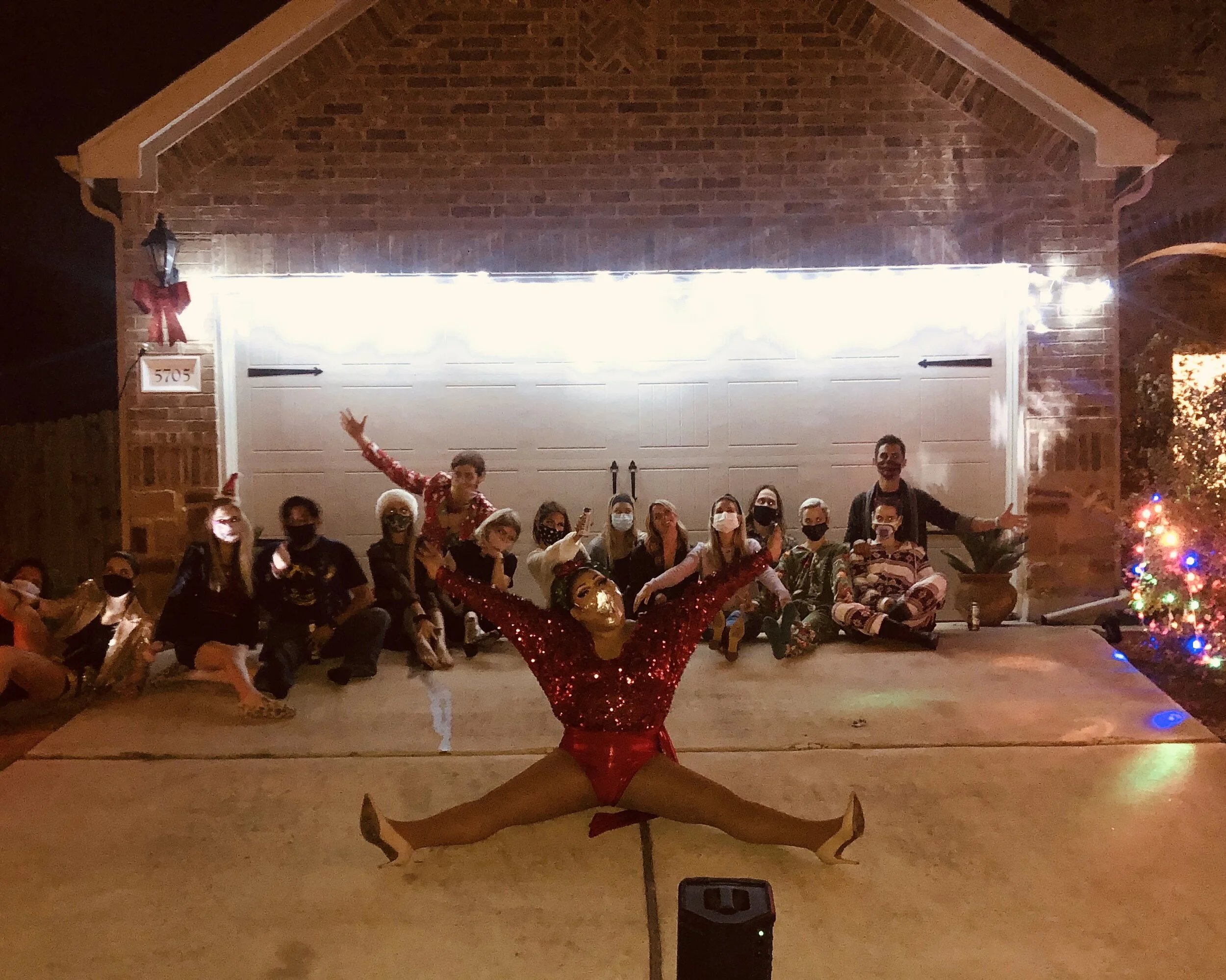 A group of people, some wearing face masks, are gathered in front of a garage door at night for a holiday celebration. In front, a dancer dressed in a shiny red costume performs a split with arms raised, a gold mask, and high heels. A decorated Chris