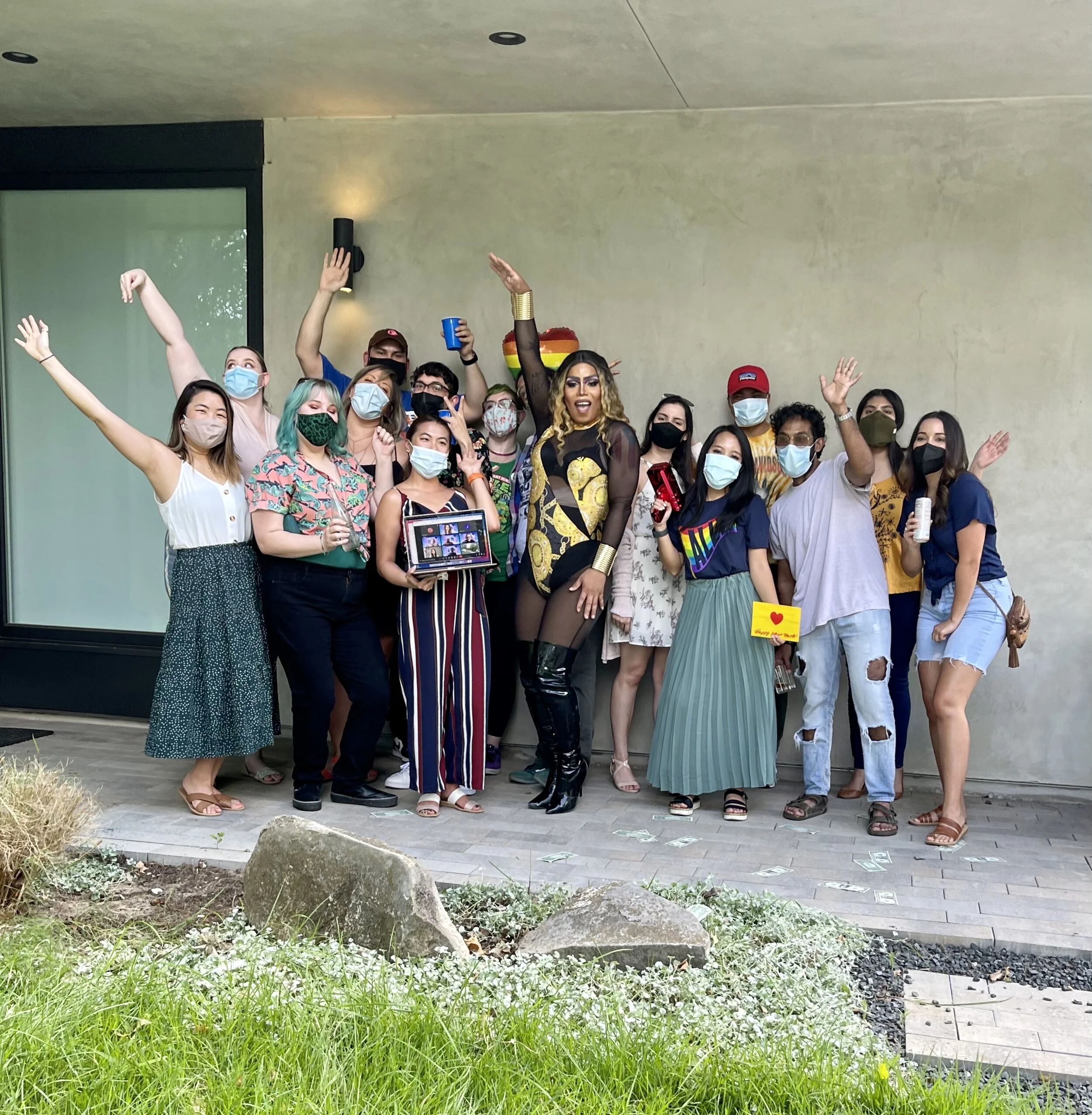 A group of people wearing face masks gathering outdoors, celebrating with a drag performer dressed in black and gold, some holding drinks and a laptop, and numerous dollar bills on the ground.