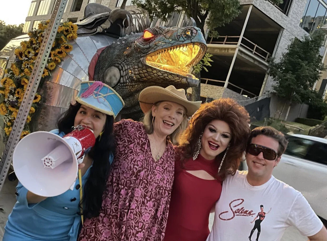 Four people posing together in front of a decorative alligator sculpture, with one person wearing a colorful hat and holding a megaphone, during a festive outdoor event.