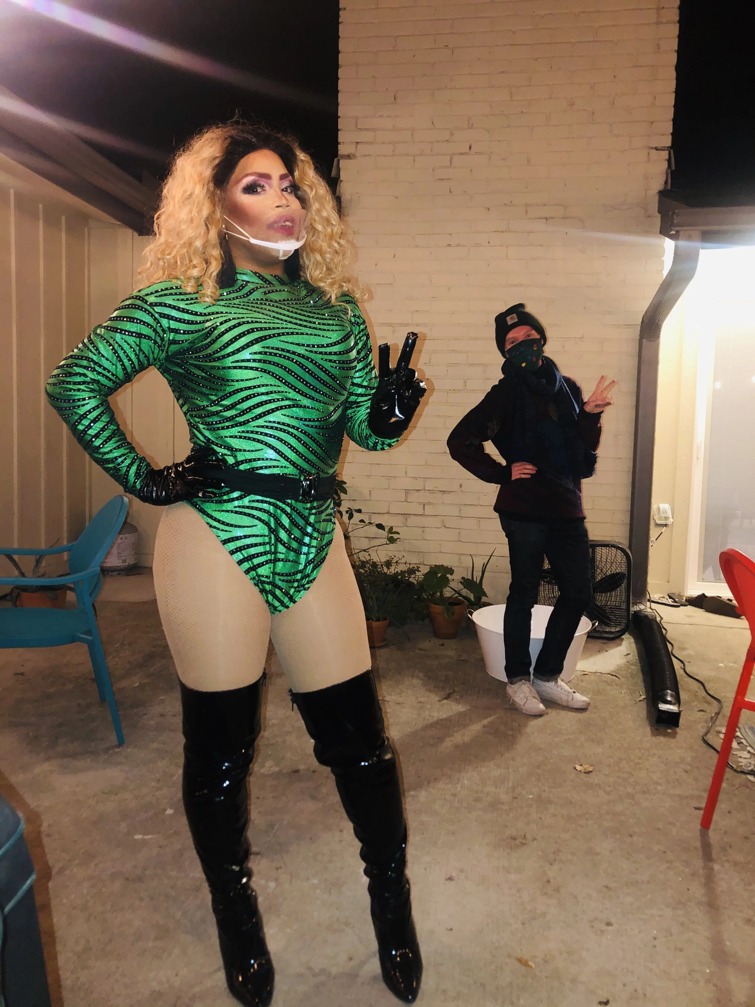 Person with curly blonde hair wearing a green zebra-striped outfit, black gloves, and black thigh-high boots, posing with a peace sign. Someone in the background is dressed in black with a beanie and face covering, standing outdoors at night near a b