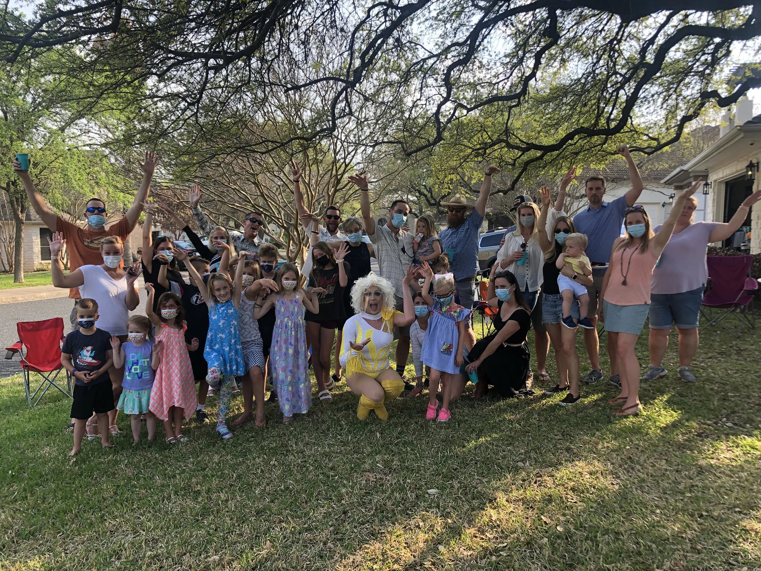 Group of people, including children and adults, gathered outdoors in a park-like area with trees, celebrating together. Some are wearing face masks and casual summer clothes, with one person dressed as a clown or performer with blond hair and colorfu