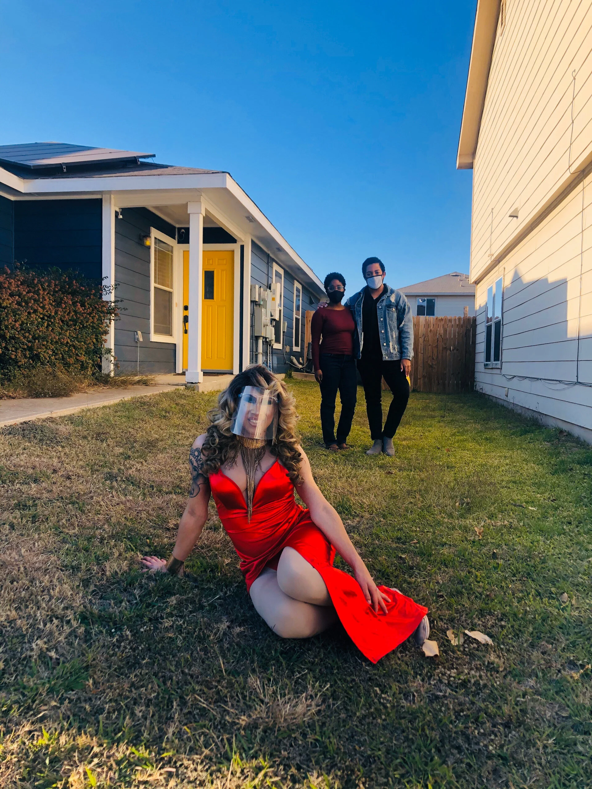 A woman in a red dress and face shield sitting on grass in front of two people standing, all wearing face masks, in a backyard with houses and a clear blue sky.