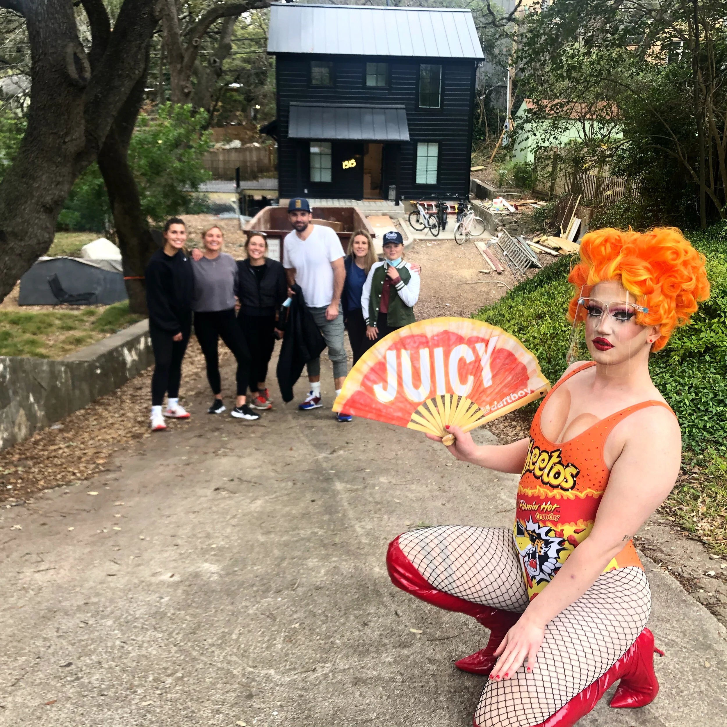A person dressed as a flamboyant drag queen with bright orange curly hair, red makeup, fishnet stockings, bright red boots, and a cow print tank top, is kneeling on the ground holding a fan with the word 'JUICY' on it. Behind them, five people are st