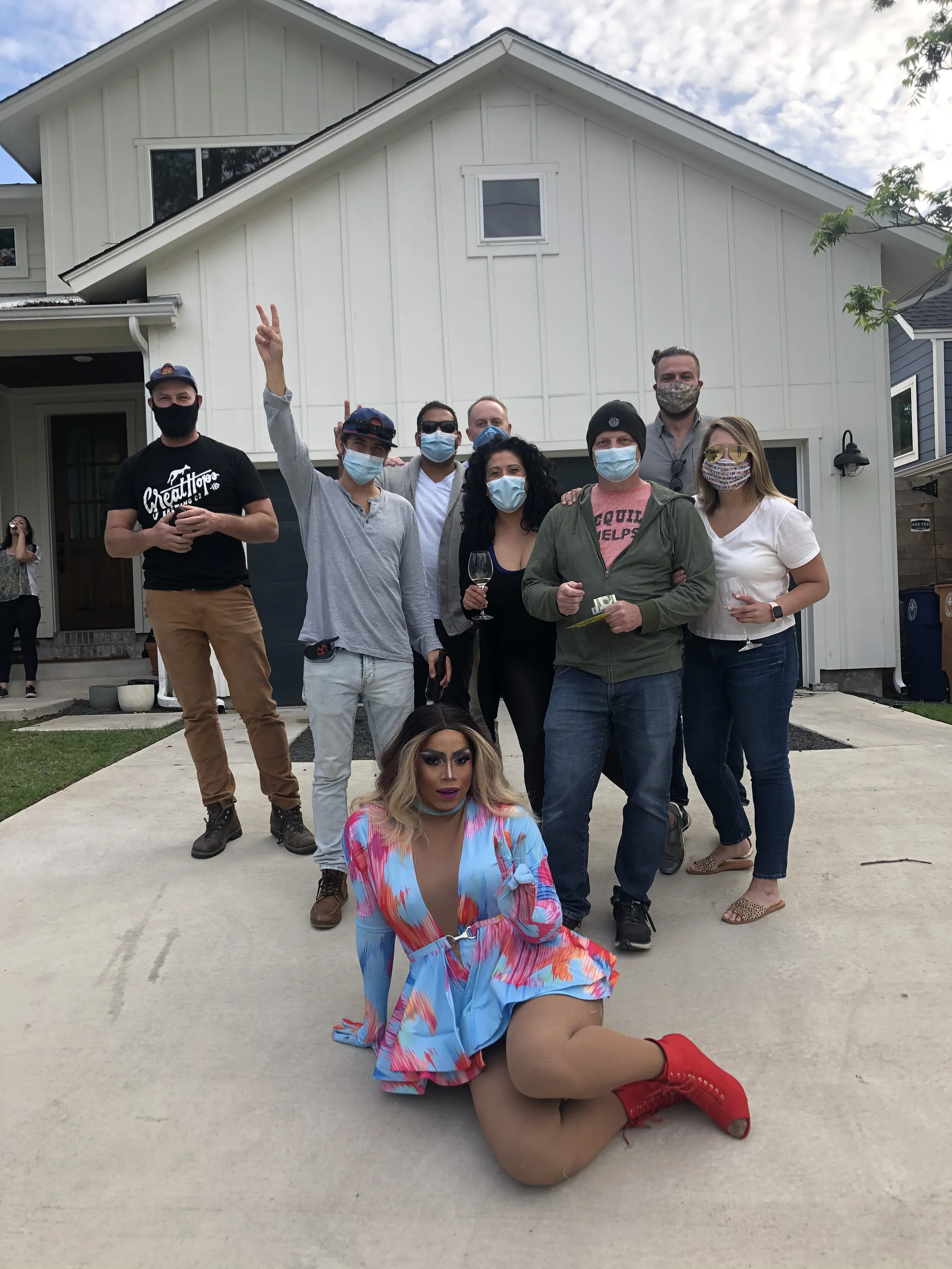 Group of people gathered in front of a house, some wearing masks, with one person sitting on the ground in colorful outfit and red boots in the foreground.