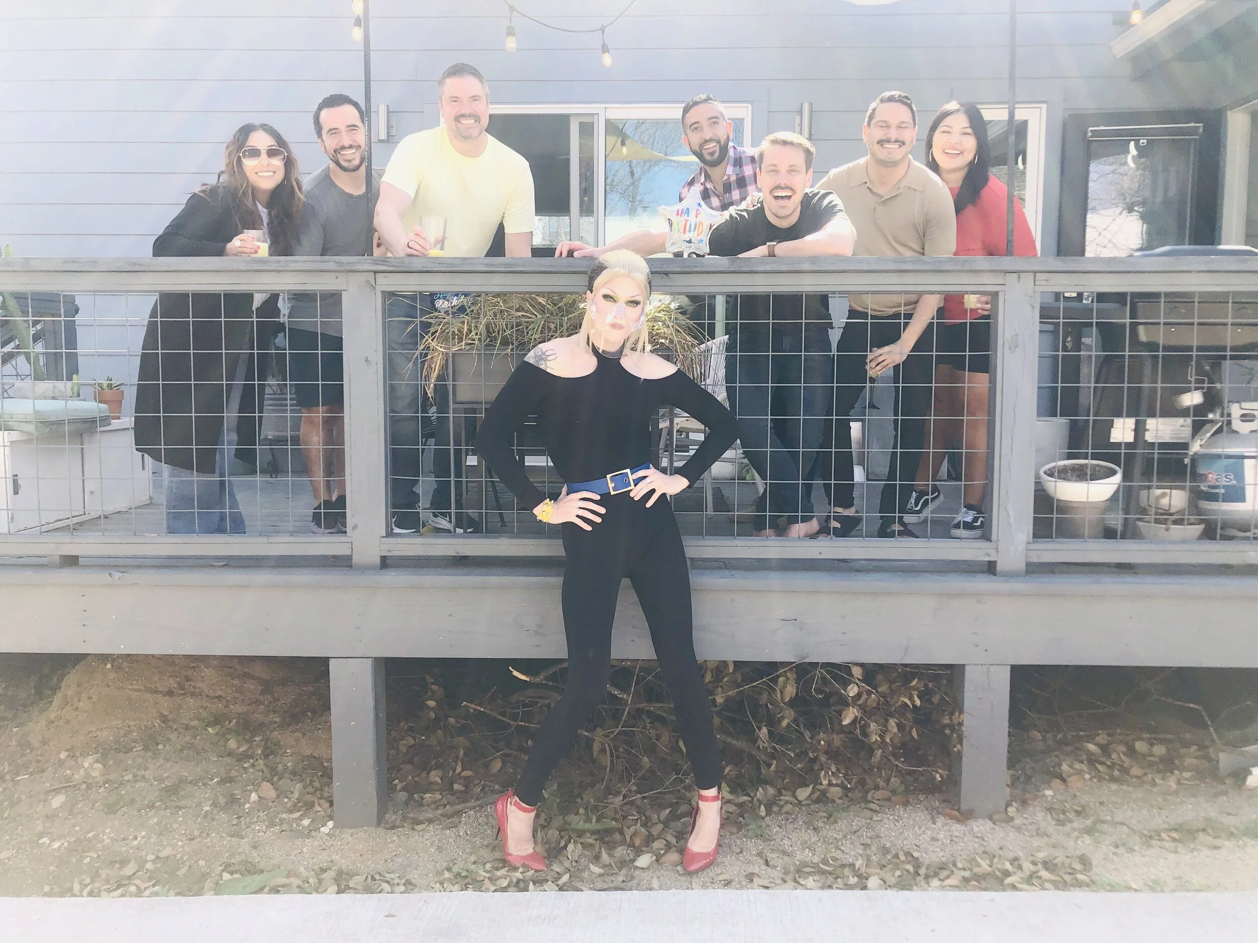 A group of nine people and one person dressed in black posing on a deck. The group stands behind a railing and smiles for the photo, while the person in black stands in front with hands on hips, wearing red high heels and bold makeup.