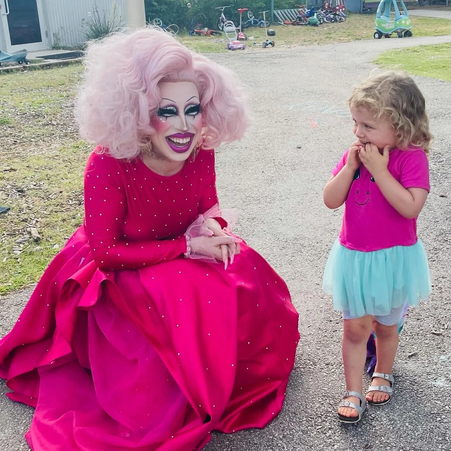 A performer dressed in a bright pink gown with dramatic makeup and voluminous pink hair crouches while talking to a young girl with curly blonde hair, wearing a pink t-shirt, a light blue tutu, and sandals, who is giggling and covering her mouth with
