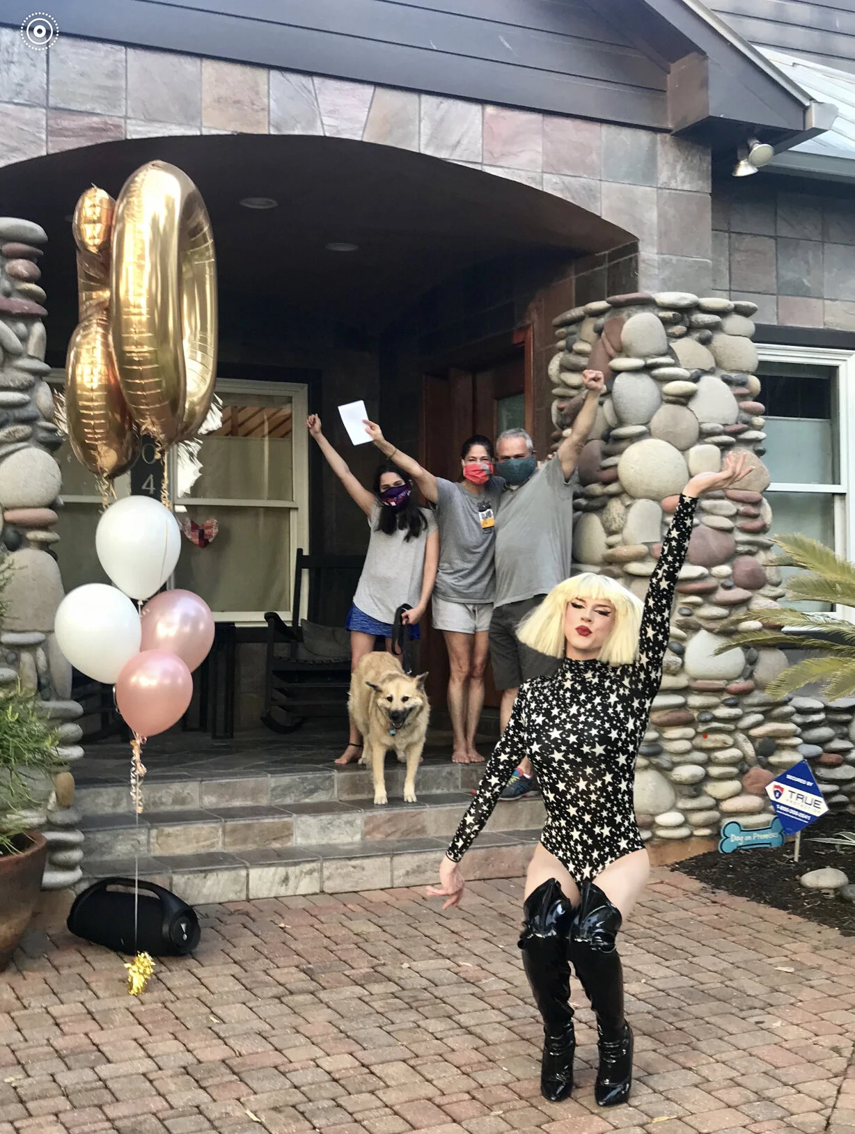 A woman with blonde hair and black star-patterned outfit posing in front of a house with a group of people and a dog on the porch celebrating, with balloons and decorations.