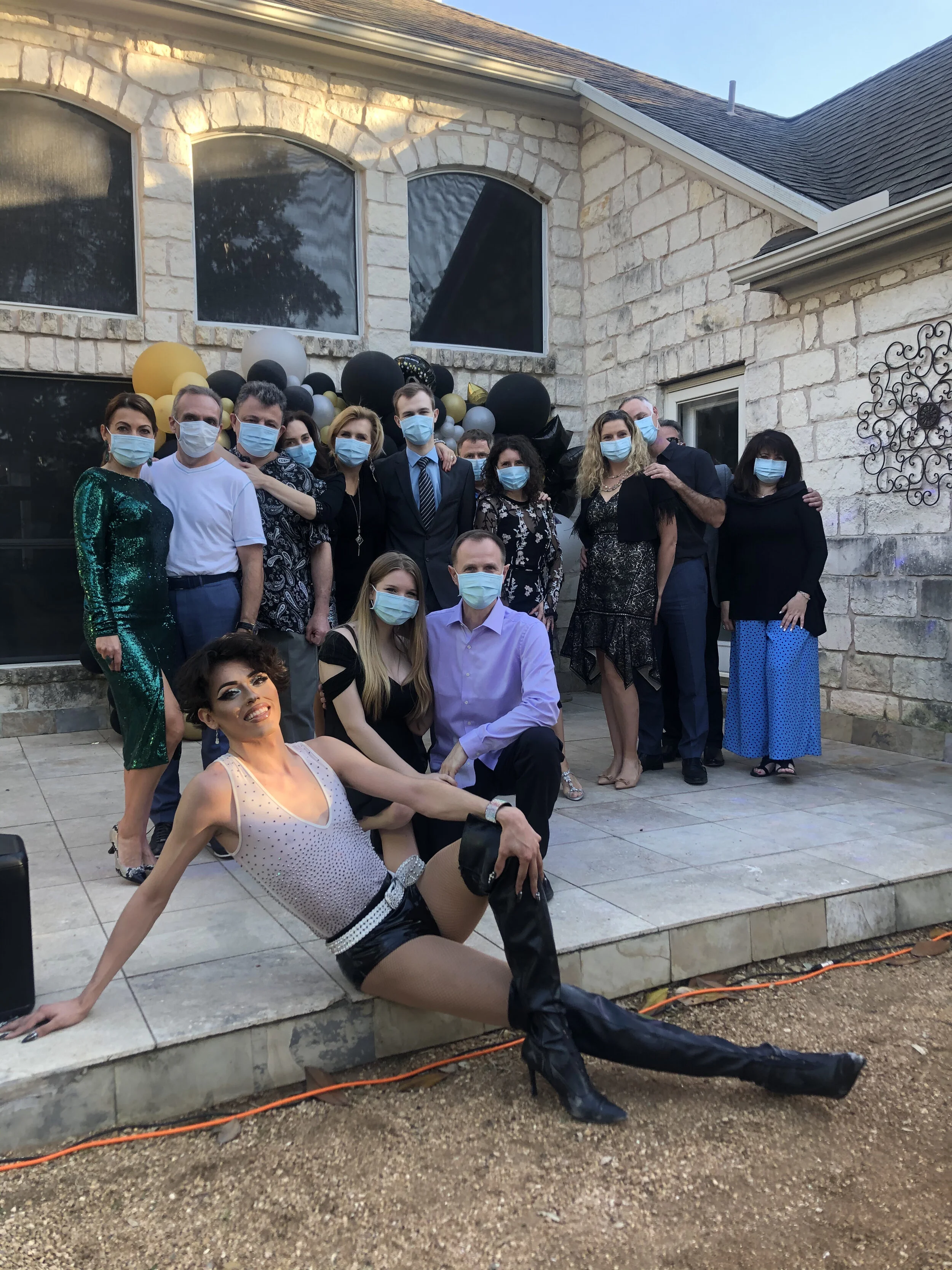 A group of people standing outside a stone house, all wearing face masks, celebrating a special occasion with black, gold, and gray balloons in the background. A woman in front is sitting on the ground with one leg extended and smiling at the camera.