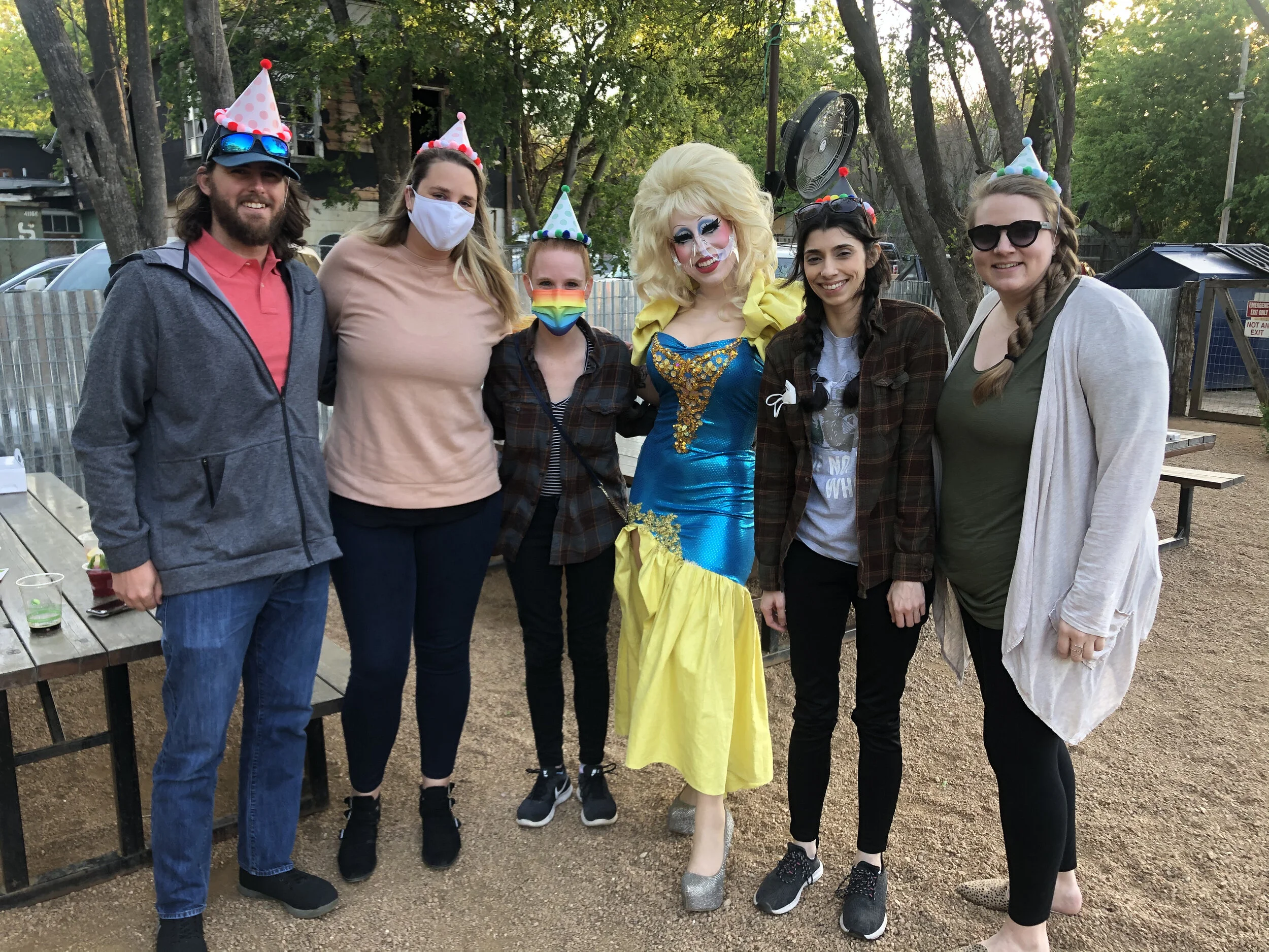 Group of six people dressed casually and celebrating at an outdoor party, with some wearing party hats and face masks, and a person in a colorful princess costume in the center.