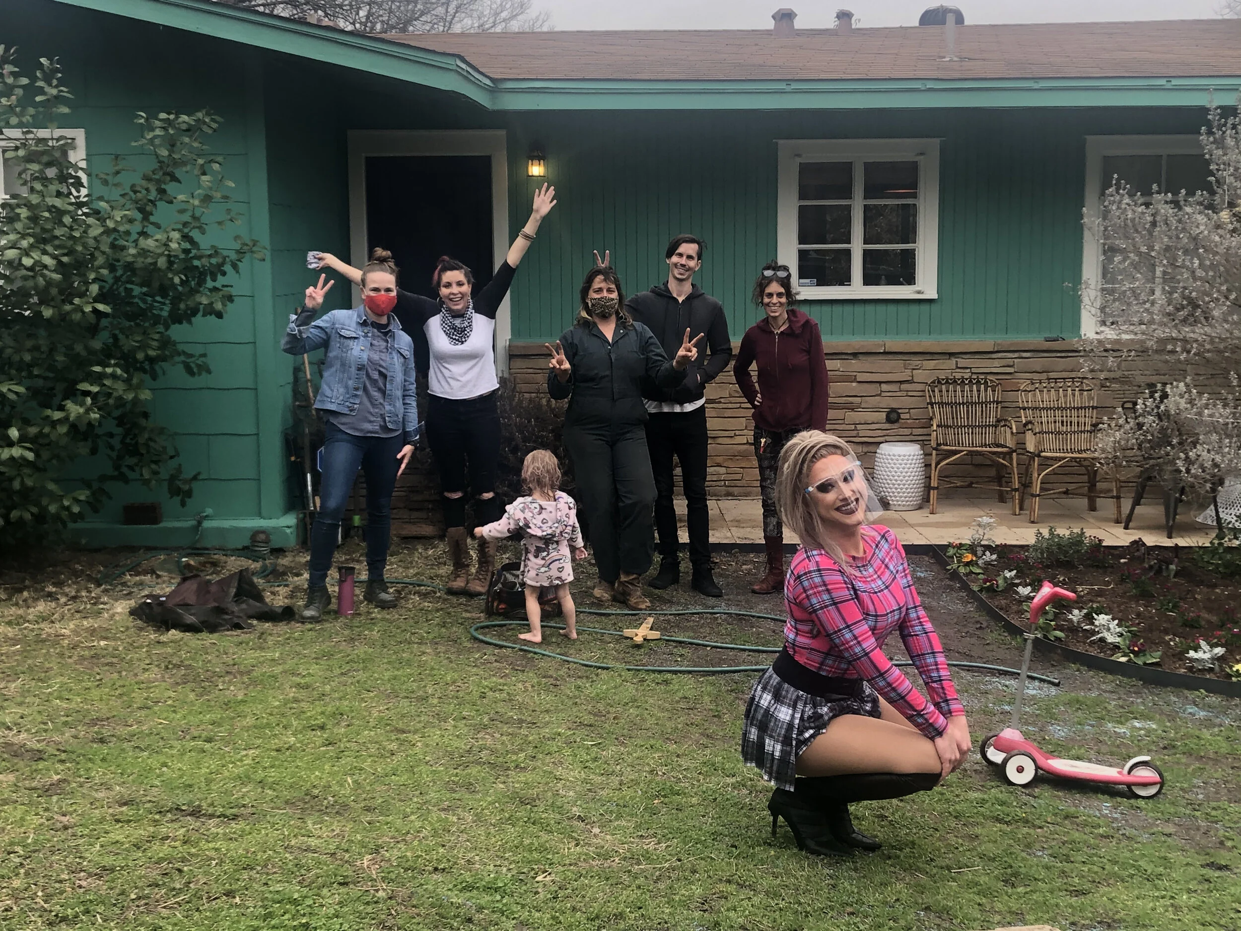 Group of people standing and smiling in a backyard, with a woman in a plaid shirt and high heels kneeling in the foreground near a pink scooter on the grass.