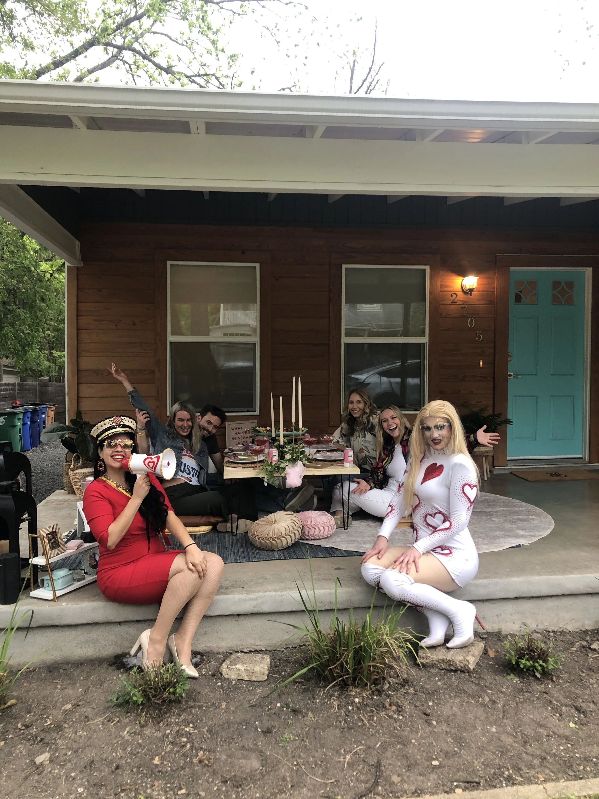 A group of seven women having a celebration outdoors on a porch, with two women dressed in distinctive outfits: one in a red dress with a megaphone and captain's hat, and another in a white dress with red hearts and knee-high boots. The table has bir