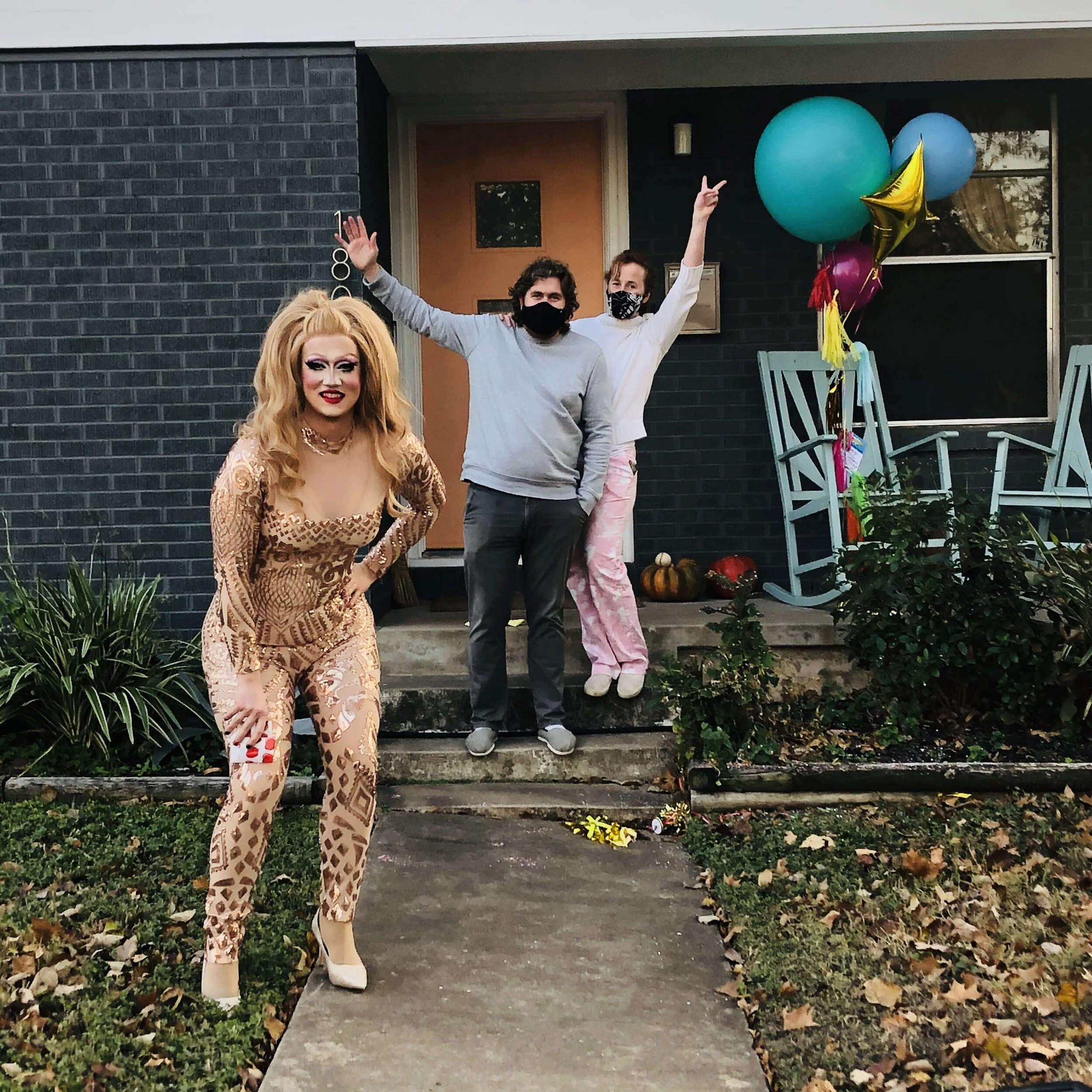 Three people standing on a front porch decorated for a celebration, with colorful balloons and pumpkins. The person on the left is dressed in a gold patterned outfit with makeup, and the other two are wearing masks and casual clothes, posing happily.