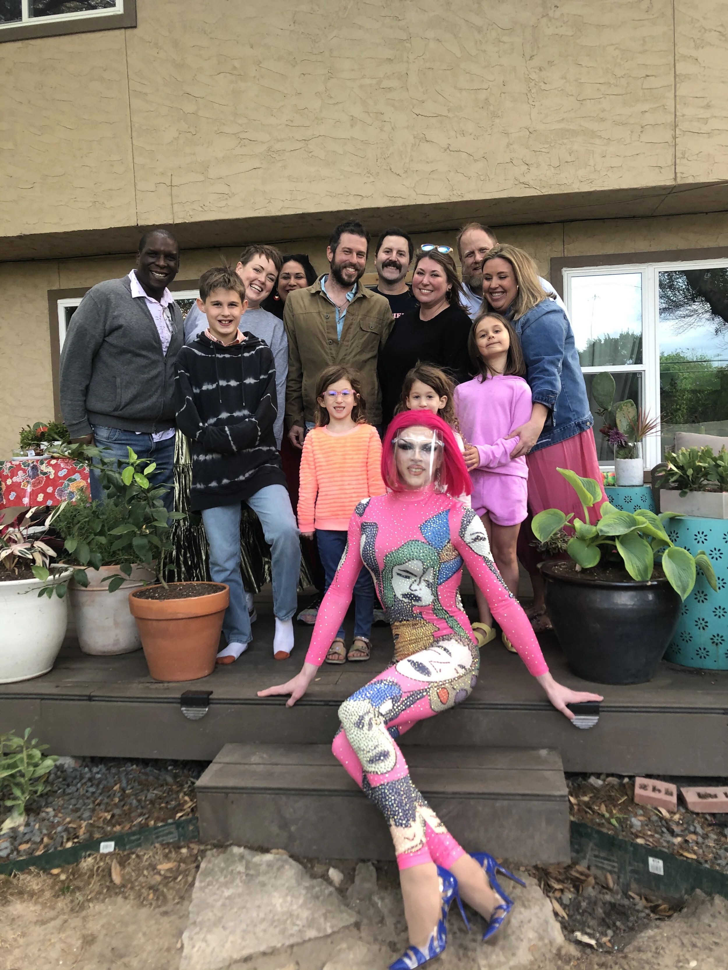 A group of people, including children and adults, posing outdoors on a wooden deck with potted plants, with a person dressed in a colorful, artistic costume sitting in front.