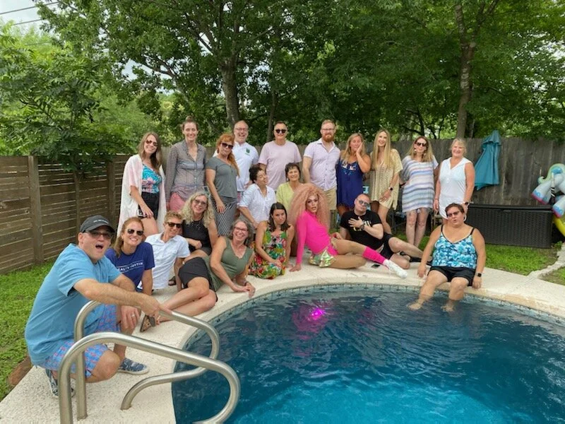 Group of people gathered around a backyard swimming pool, some sitting on the pool edge and others standing behind, wearing summer clothes and sunglasses, with trees and a wooden fence in the background.
