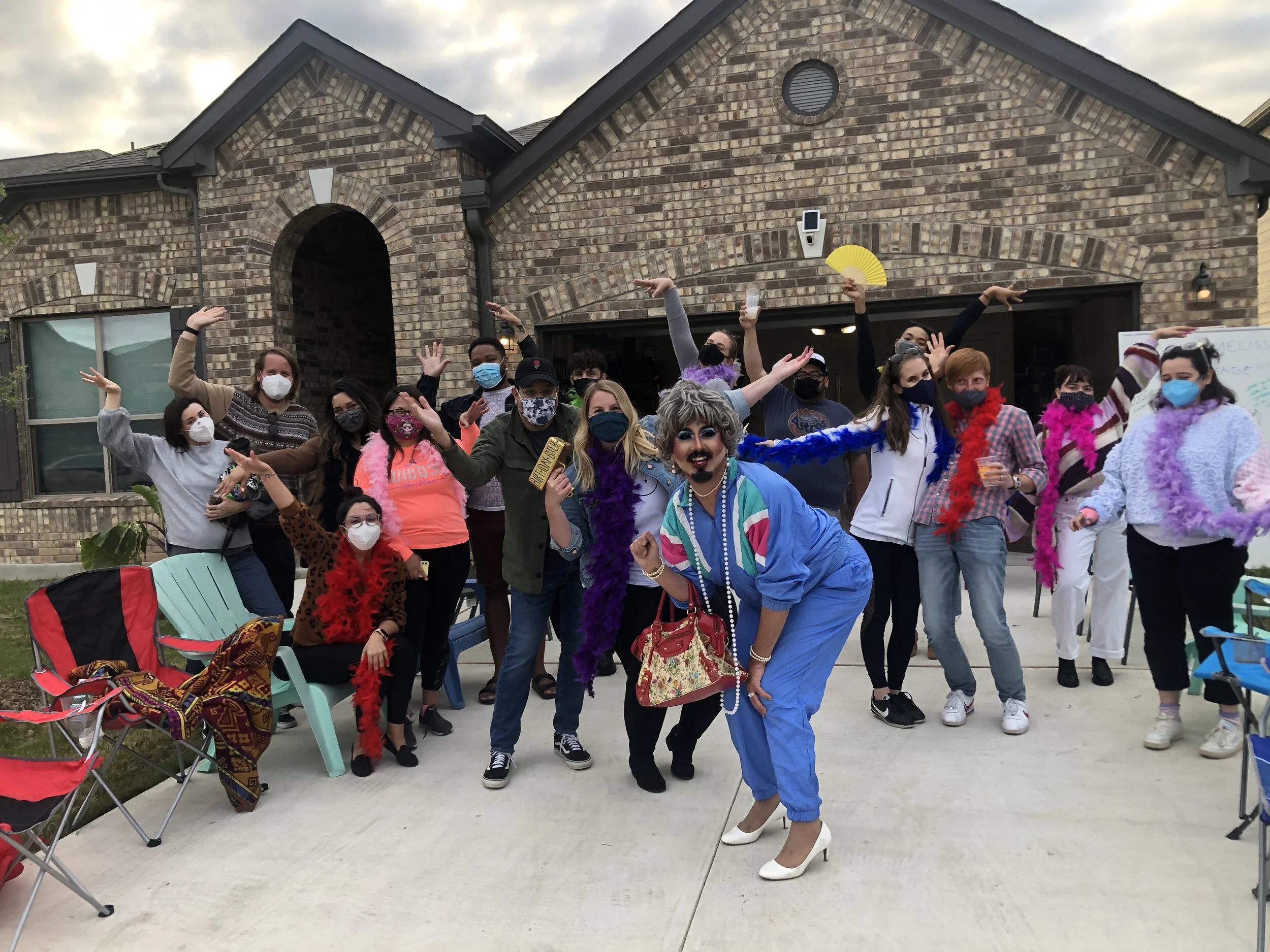 Group of diverse people celebrating outdoors, wearing colorful costumes and masks, with some holding props like fans and gold bars, in front of a brick house during evening.