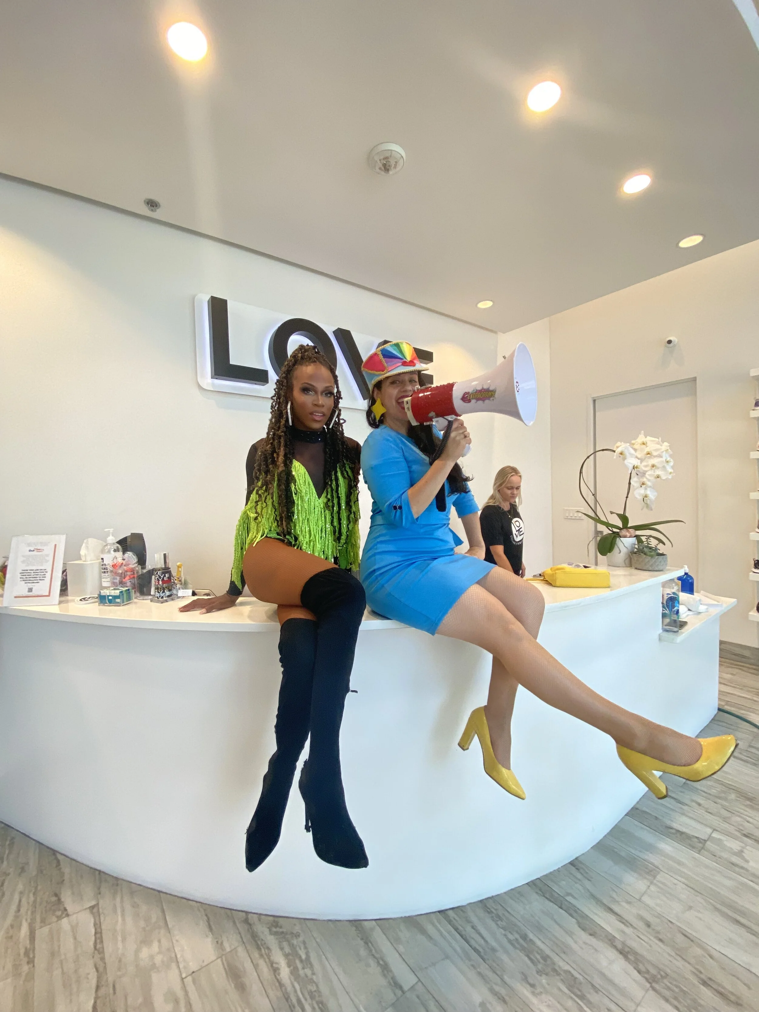 Two women sitting on a white reception desk; one wearing lime green fringe top and black thigh-high boots, the other in a blue dress with yellow heels, holding a megaphone, with the word 'LOVE' on the wall behind them.